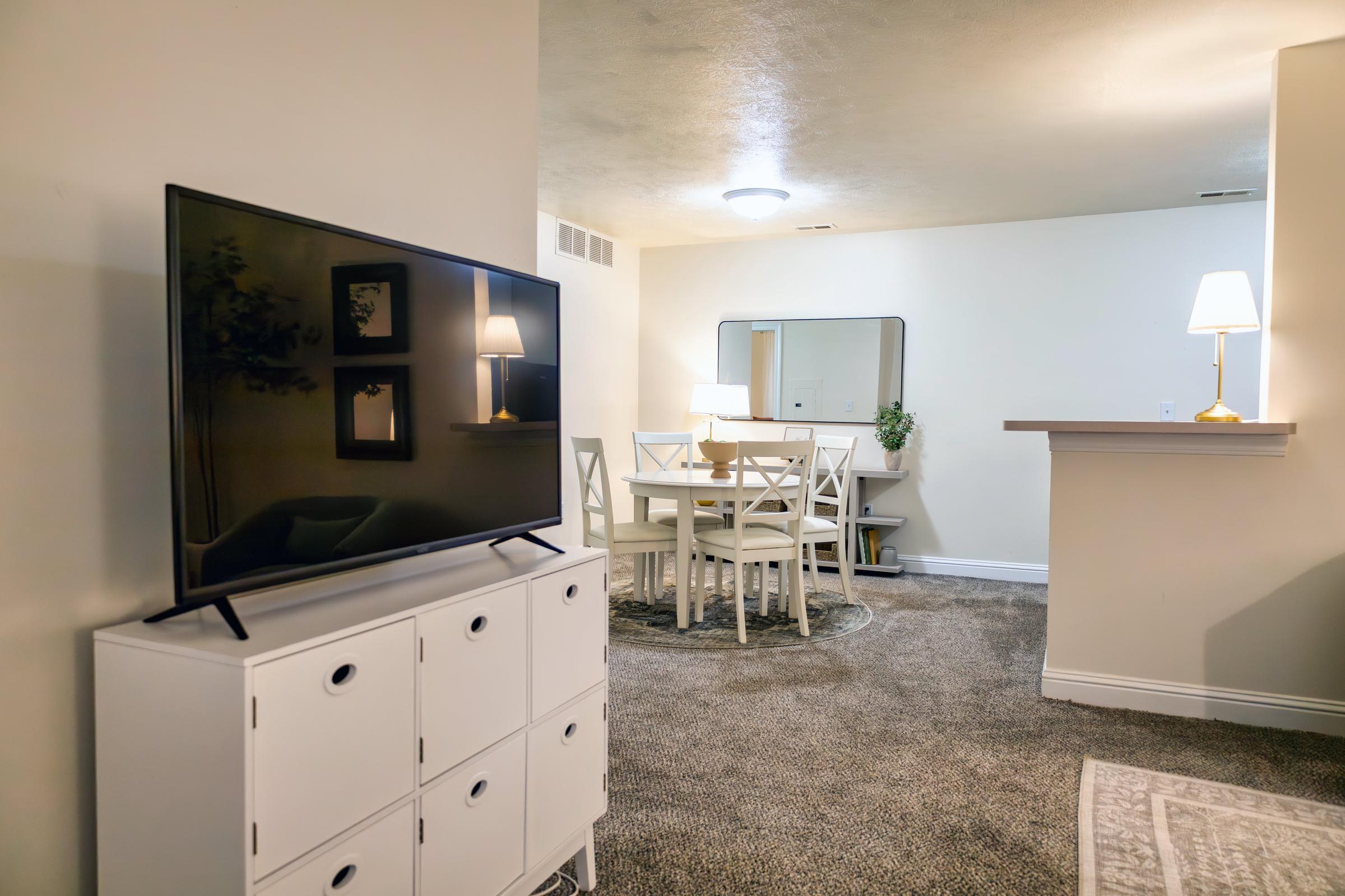 A cozy living space featuring a large television on a white media console. In the background, there is a dining area with a round table, four chairs, a mirror on the wall, and a lamp providing soft lighting. The flooring is carpeted with neutral tones, and decorative elements add warmth to the room.
