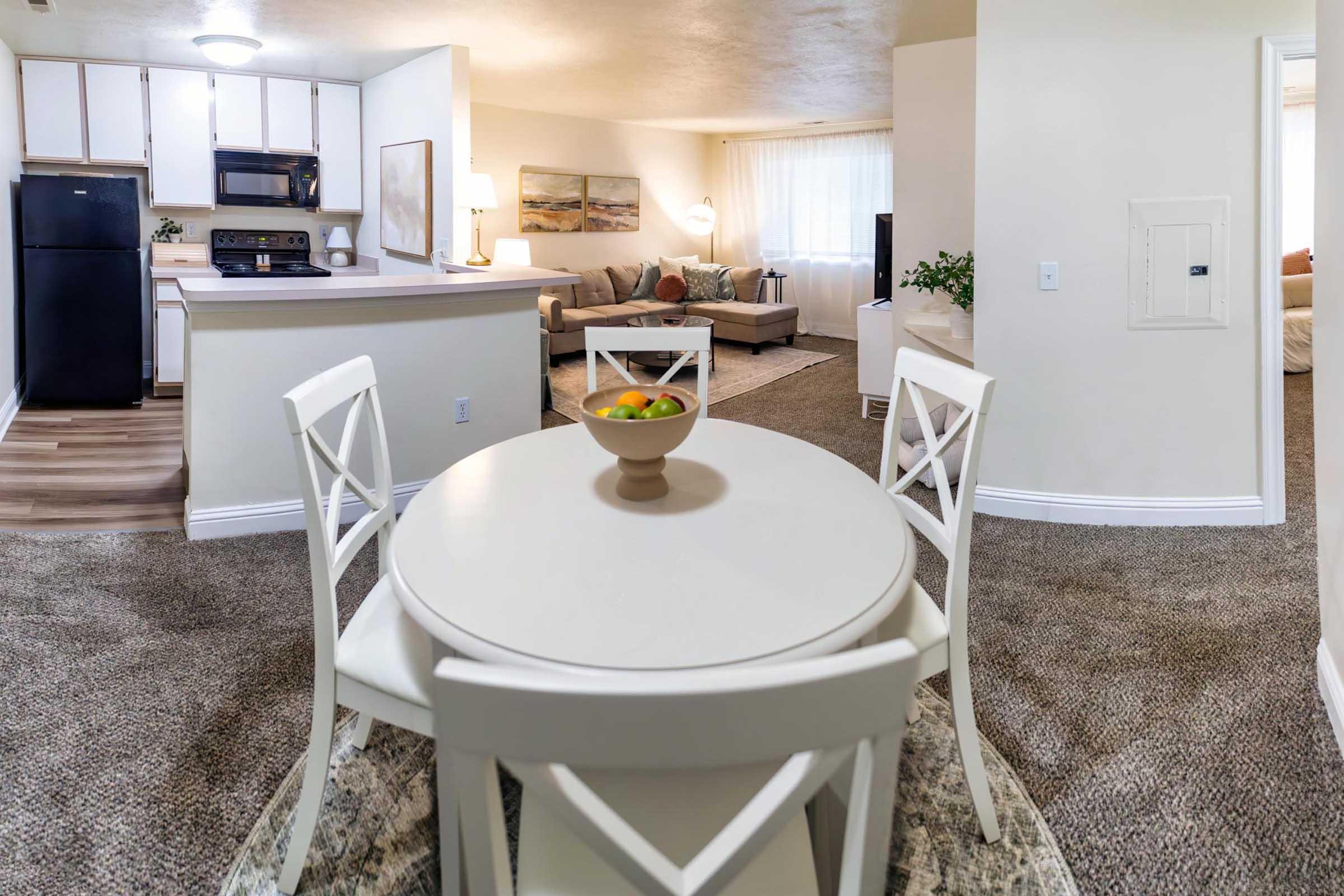 A spacious, well-lit living area featuring a kitchen with black appliances, a small dining table with four chairs, and a cozy seating area with a beige couch. The room includes decorative items and a bowl of fruit on the table, creating a welcoming atmosphere. Soft natural light filters through the curtains.
