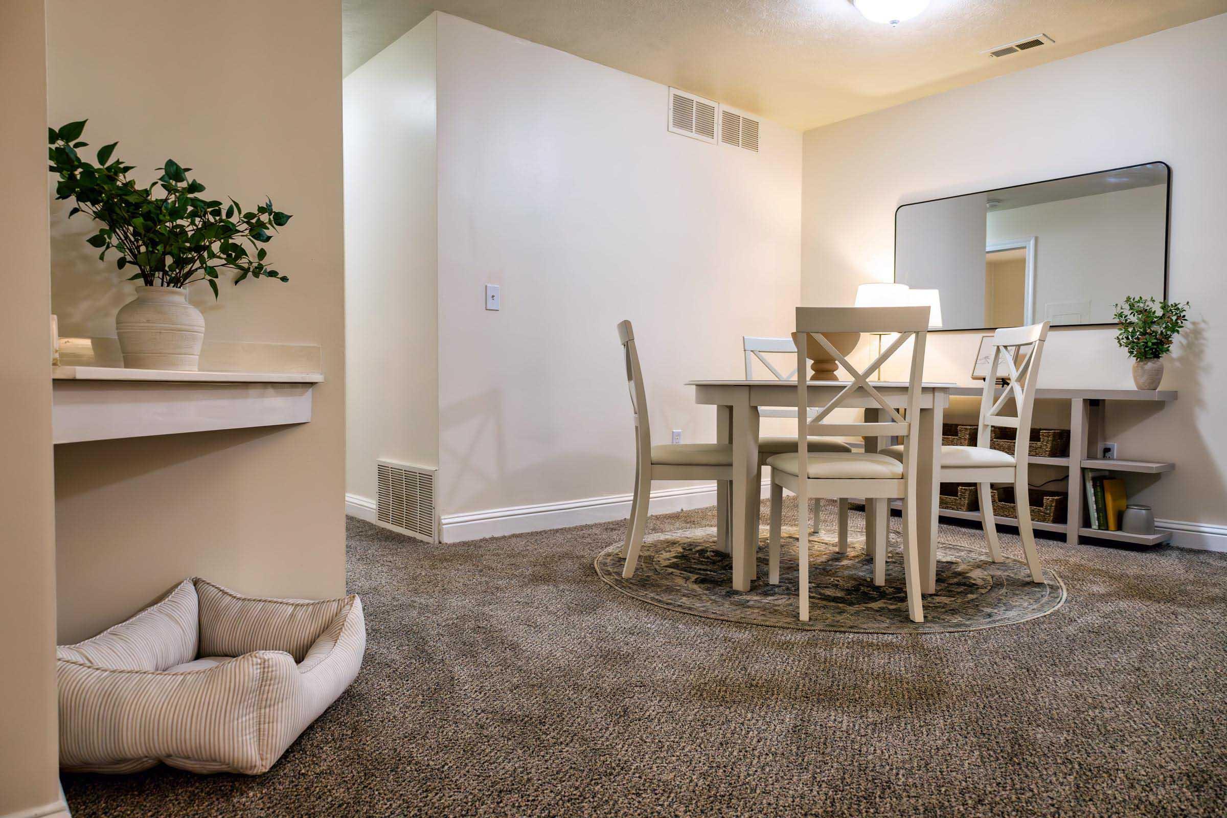 A cozy dining area featuring a round table surrounded by four chairs, a decorative mirror on the wall, and a soft rug. A plant in a minimalist vase adds a touch of greenery, while a pet bed sits on the carpeted floor nearby, creating a warm and inviting atmosphere.