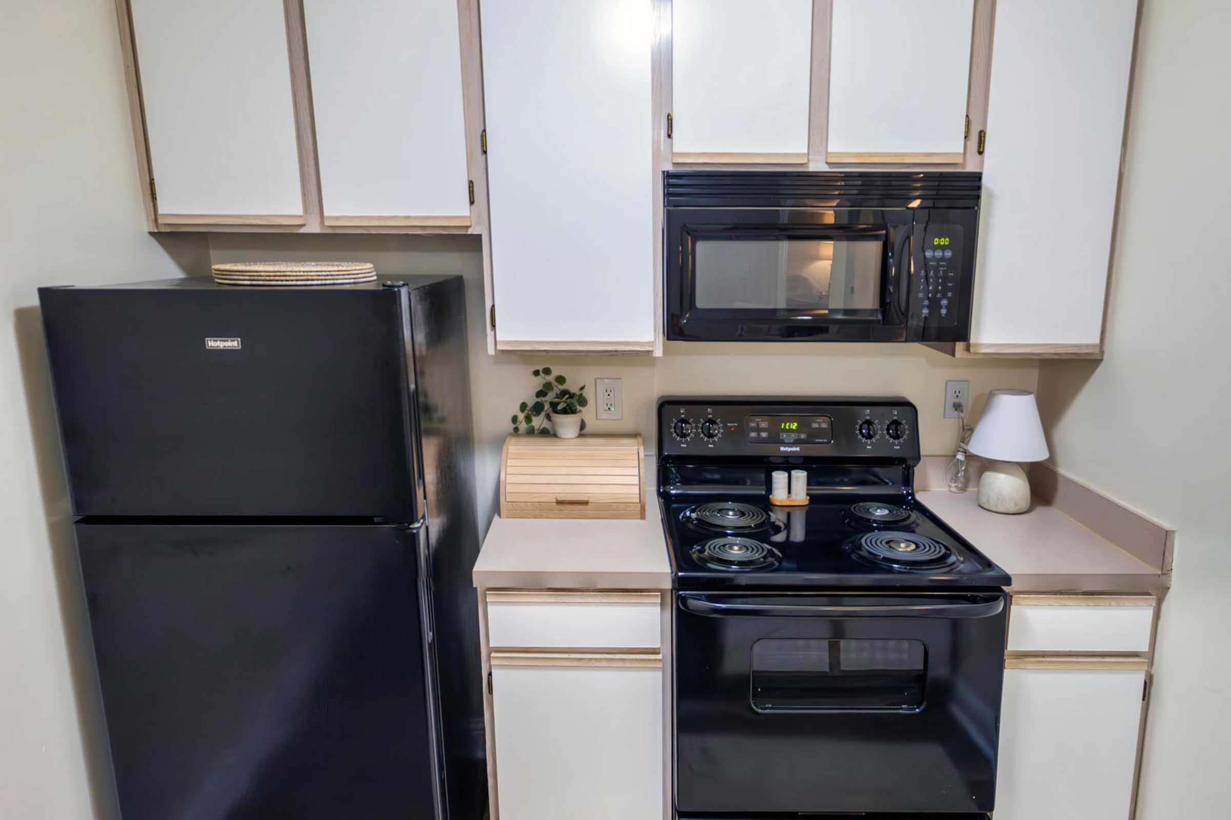 A kitchen featuring a black refrigerator, a black stove with four burners, and a microwave above it. Counters are light-colored, and there's a small lamp and a wooden organizer on the counter. Cabinets above the counters have a simple design with a mix of white and light wood.