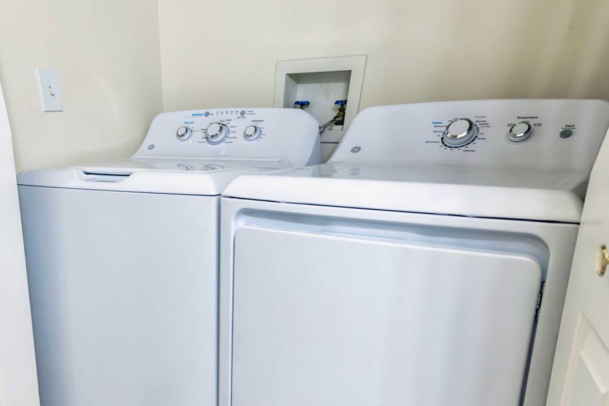 A stacked washing machine and dryer in a laundry room, both white and modern in design. The washing machine features a top-loading design, while the dryer has a front-loading appearance. They are positioned side by side against a light-colored wall.