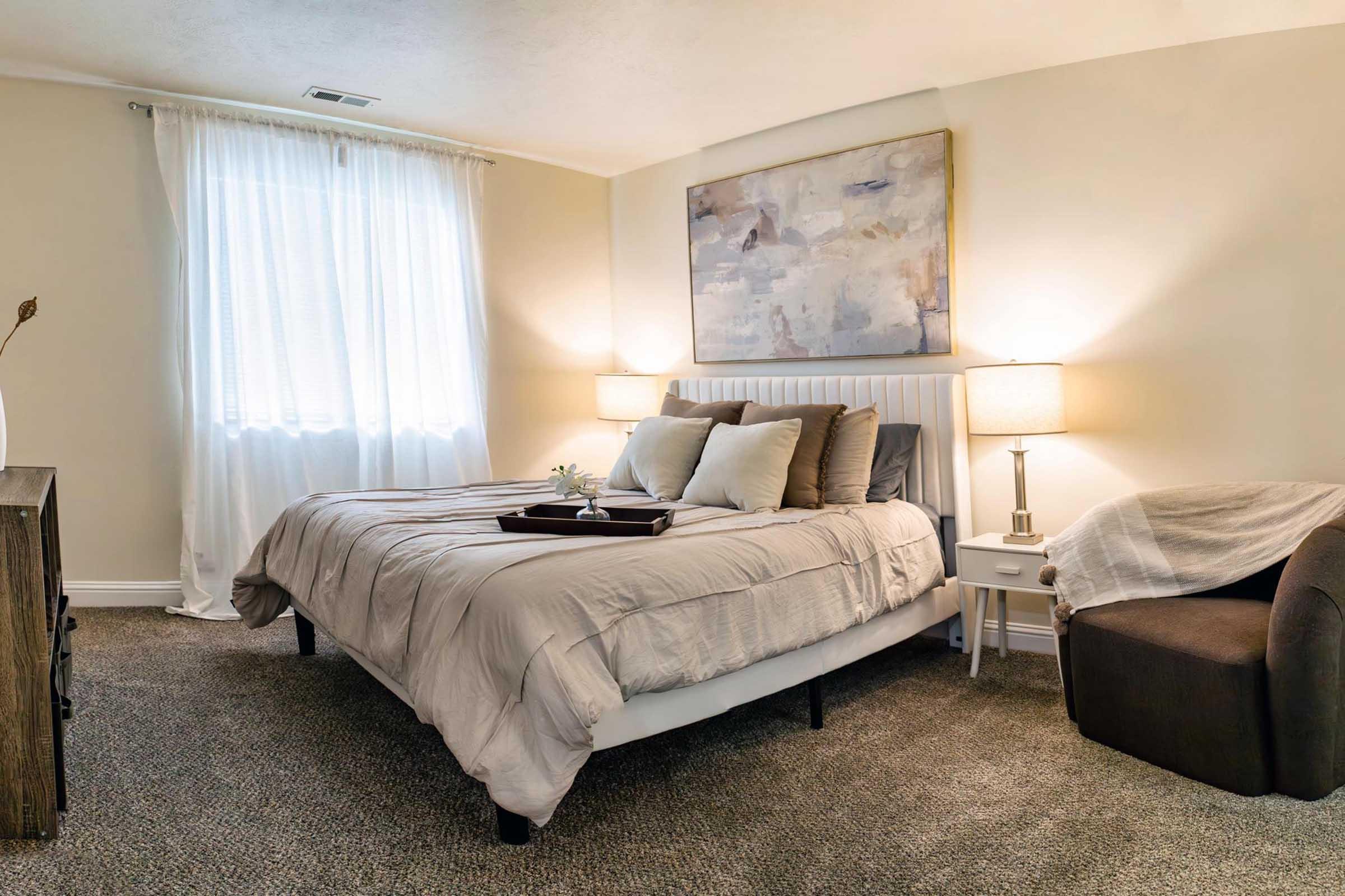 A cozy bedroom featuring a neatly made bed with neutral-colored bedding, two bedside lamps, and a wall-mounted artwork. Natural light filters through a sheer curtain. A small side table holds a decorative tray, while a plush armchair adds comfort to the space. The carpet is soft and inviting.