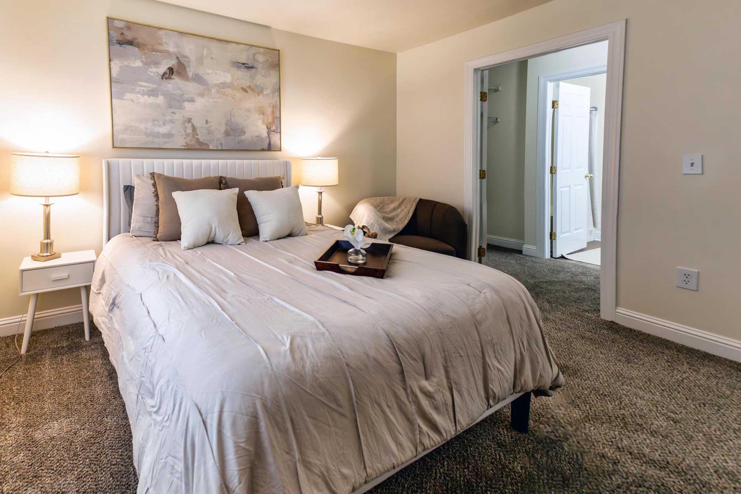 A cozy bedroom featuring a queen-sized bed with light bedding and decorative pillows. Two bedside lamps illuminate the space, which includes a small tray on the bed. A comfortable armchair is located in the corner, and a doorway leads to another room, suggesting a welcoming and well-lit atmosphere.