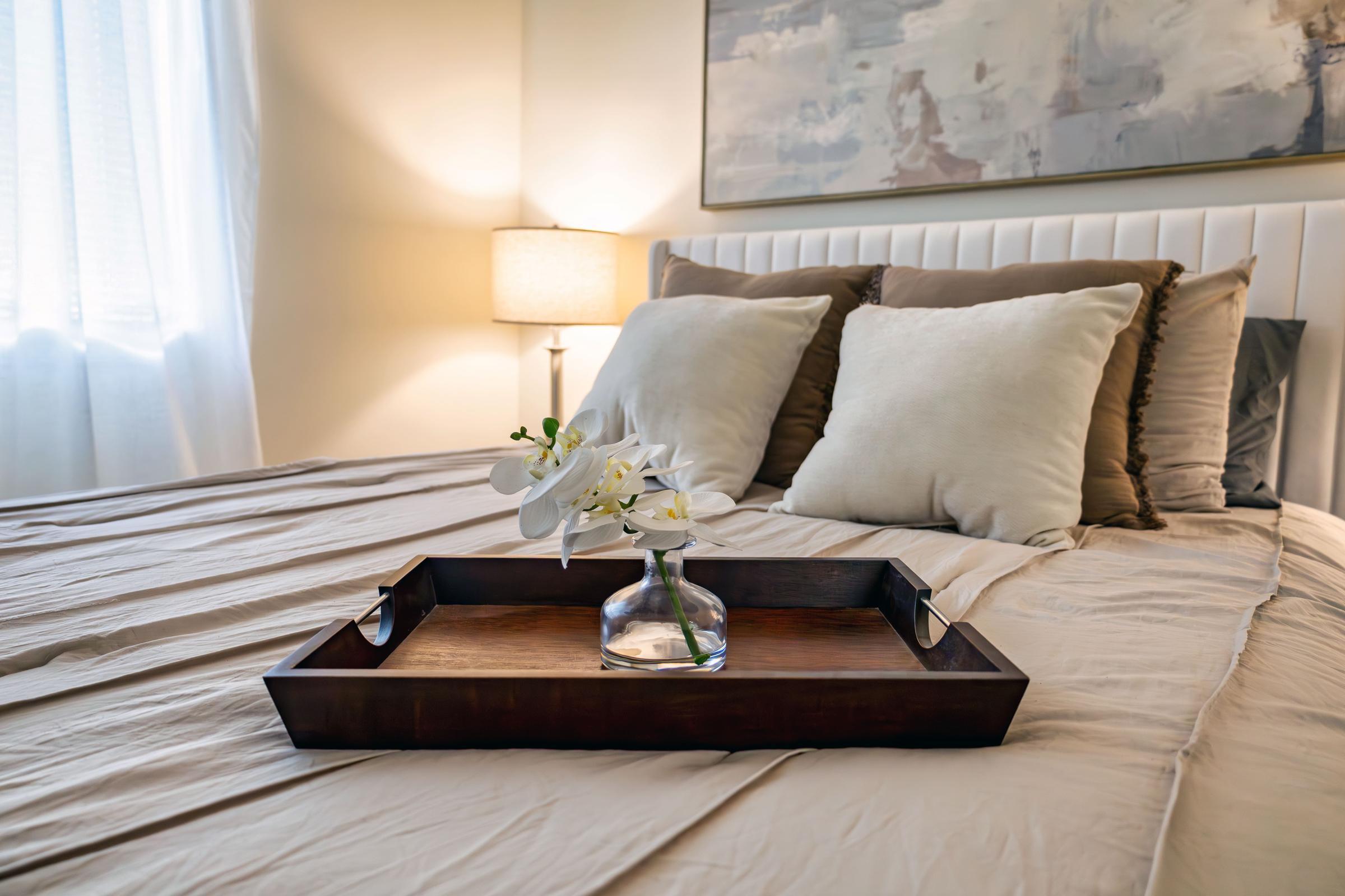 A neatly made bed with light-colored bedding, featuring two decorative pillows and a wooden tray holding a small vase of white flowers. A soft lamp illuminates the room, creating a cozy and inviting atmosphere. Abstract wall art in soft tones is visible in the background.