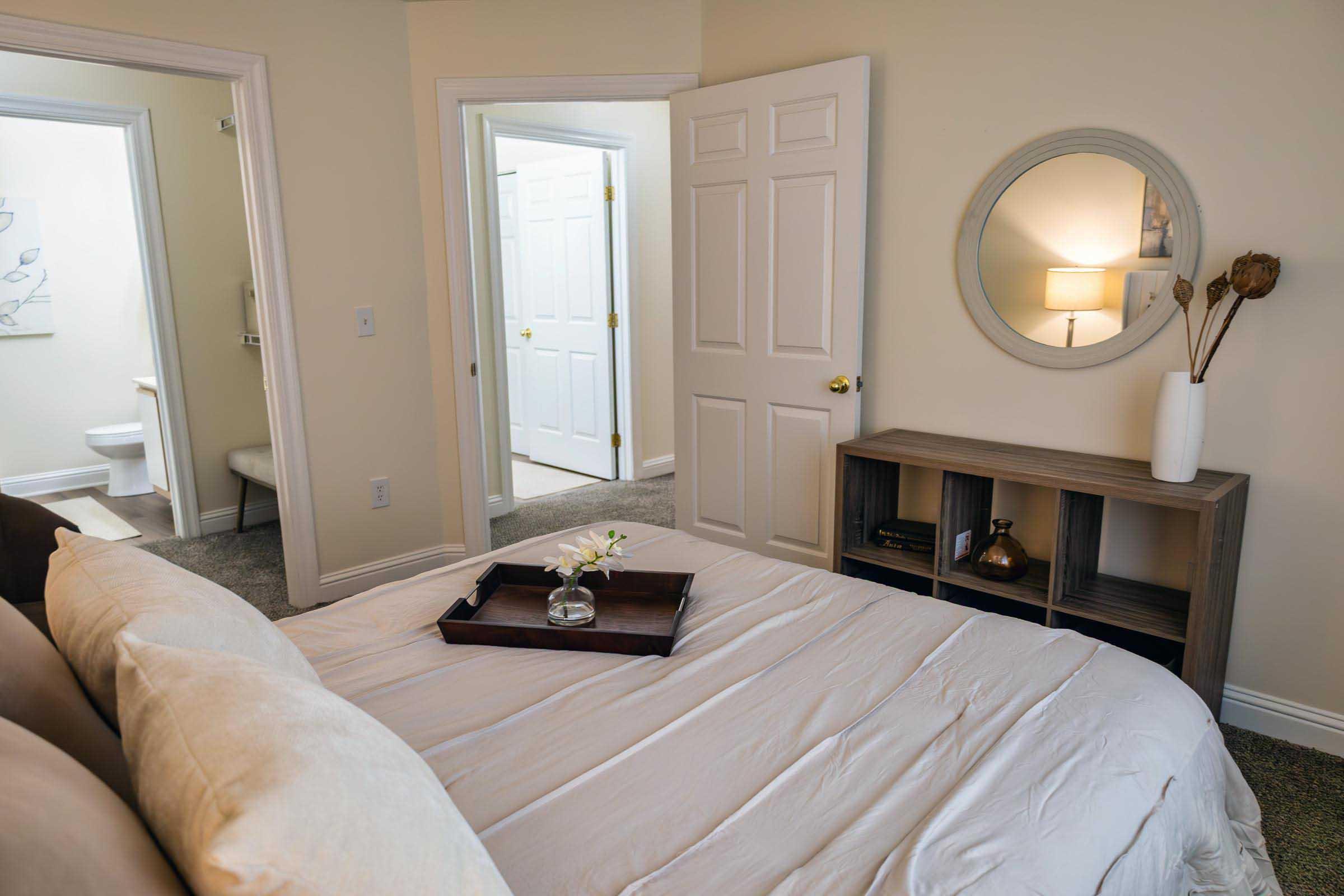 A cozy bedroom featuring a neatly made bed with light-colored bedding, a wooden nightstand with a decorative tray, and a circular mirror on the wall. The room has soft, neutral tones and a calming atmosphere, with an open door leading to a bathroom and another room visible in the background.