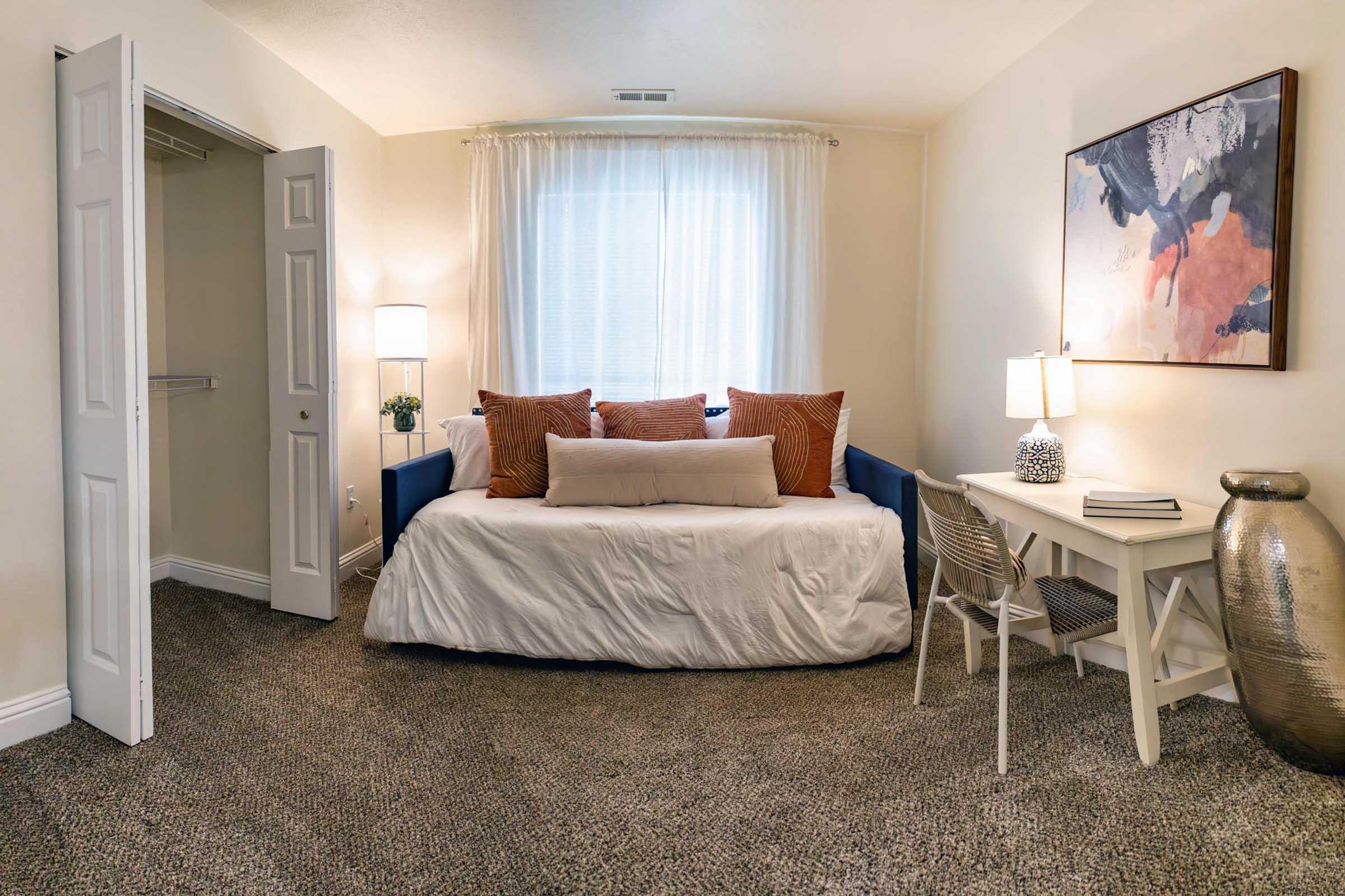 A cozy, well-lit bedroom featuring a daybed with decorative pillows, two bedside lamps, and a small desk with a chair. The walls are painted in soft colors, and a large abstract painting is hung above the desk. There is a closet with sliding doors visible to the left, and the floor is covered with carpet.