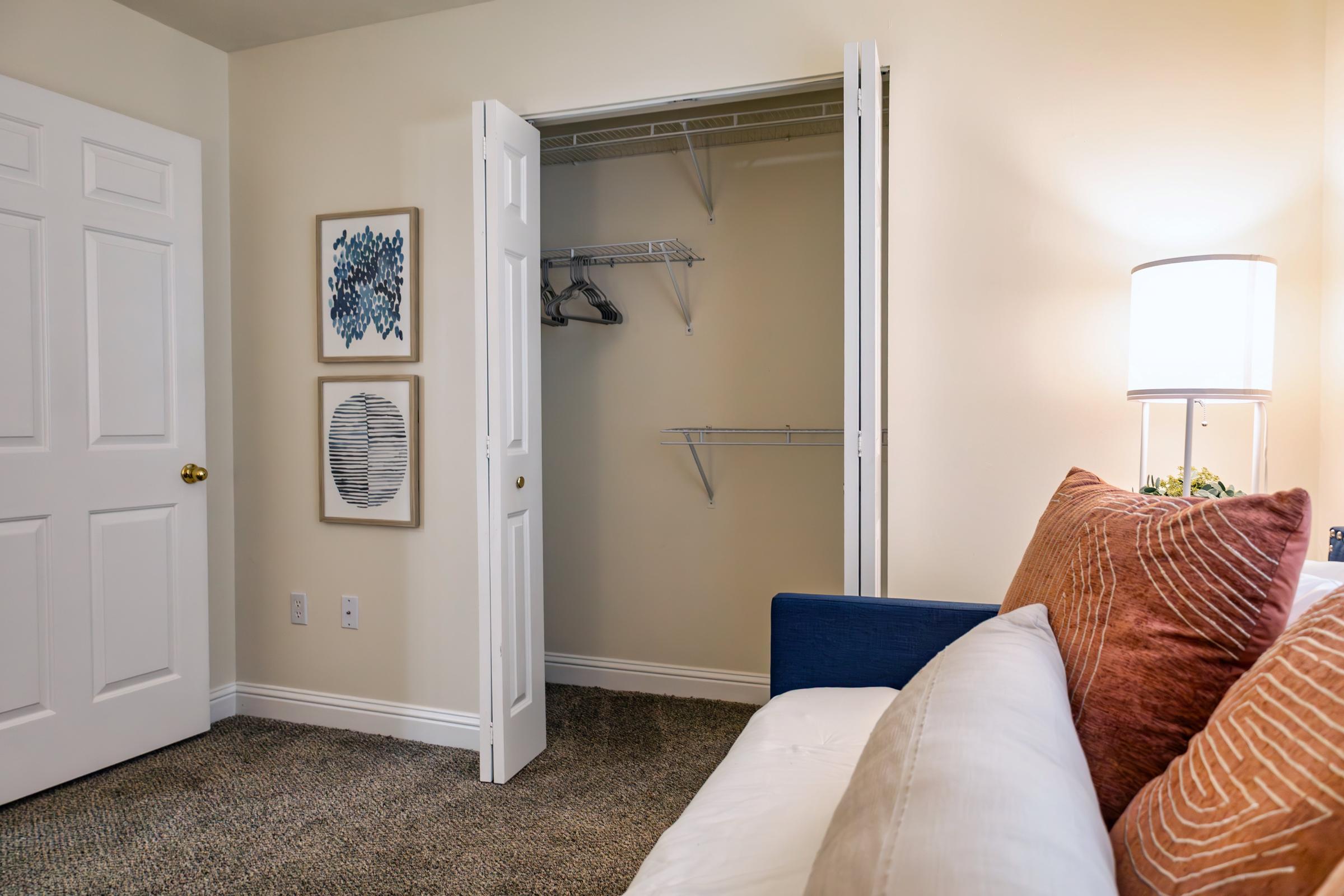 A cozy living space featuring a white sofa with orange decorative pillows, a lamp, and artwork on the wall. An open closet with shelves and hanging space is visible, showcasing a neat and organized room. The walls are painted a light color, enhancing the inviting atmosphere.