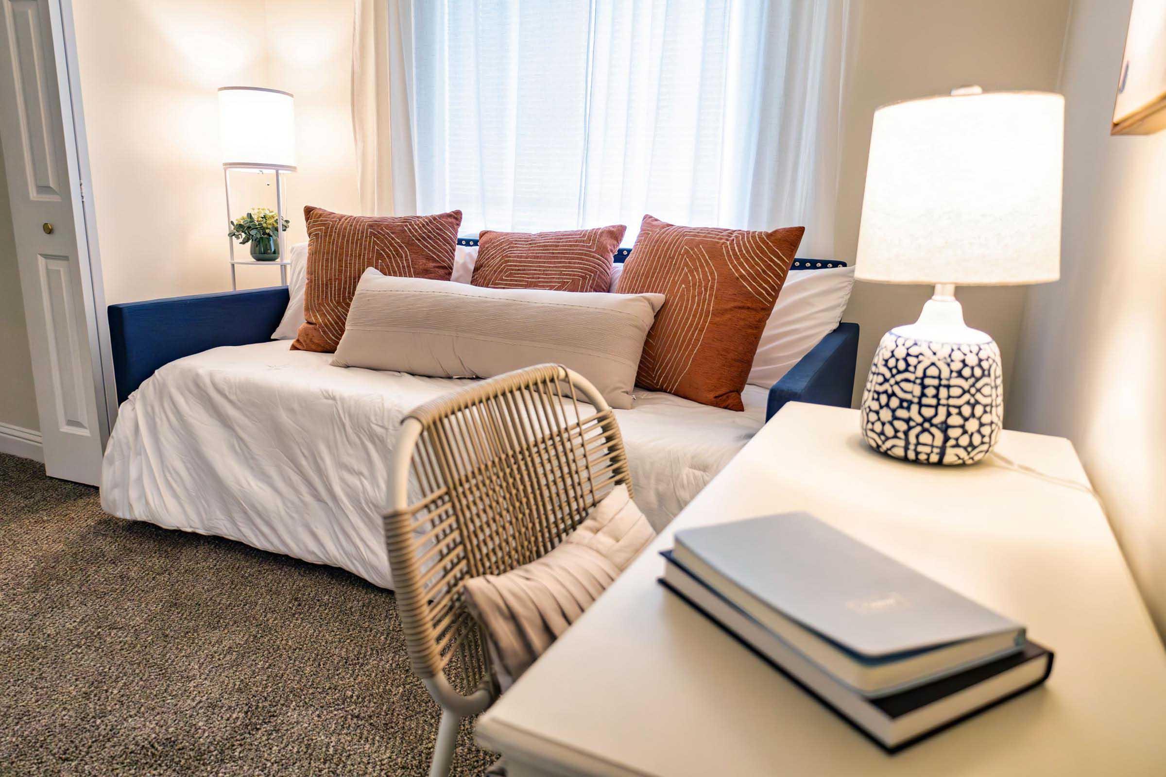 A cozy bedroom scene featuring a daybed with decorative pillows, a bedside lamp, and a small desk with a chair. Soft natural light filters through sheer curtains, creating a warm and inviting atmosphere. A stack of books rests on the desk, enhancing the room's aesthetic.