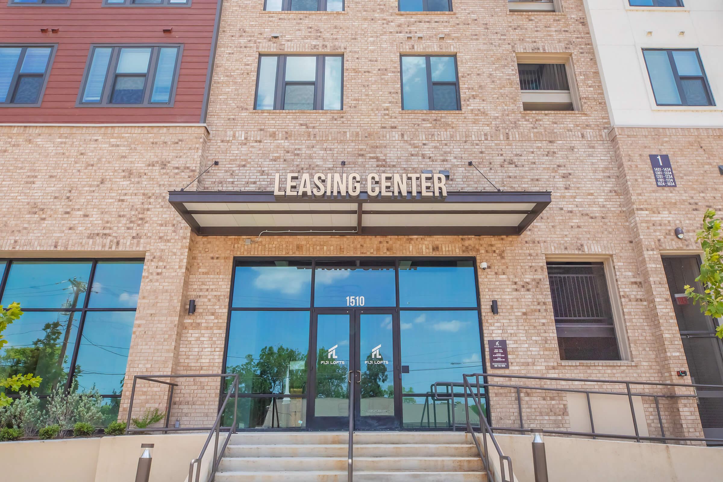 A modern leasing center with a large glass entrance surrounded by a landscaped area. The building features a mix of brick and siding in various colors. Above the door, bold letters state "LEASING CENTER," and the address "1510" is displayed prominently. The sky is blue with a few clouds.