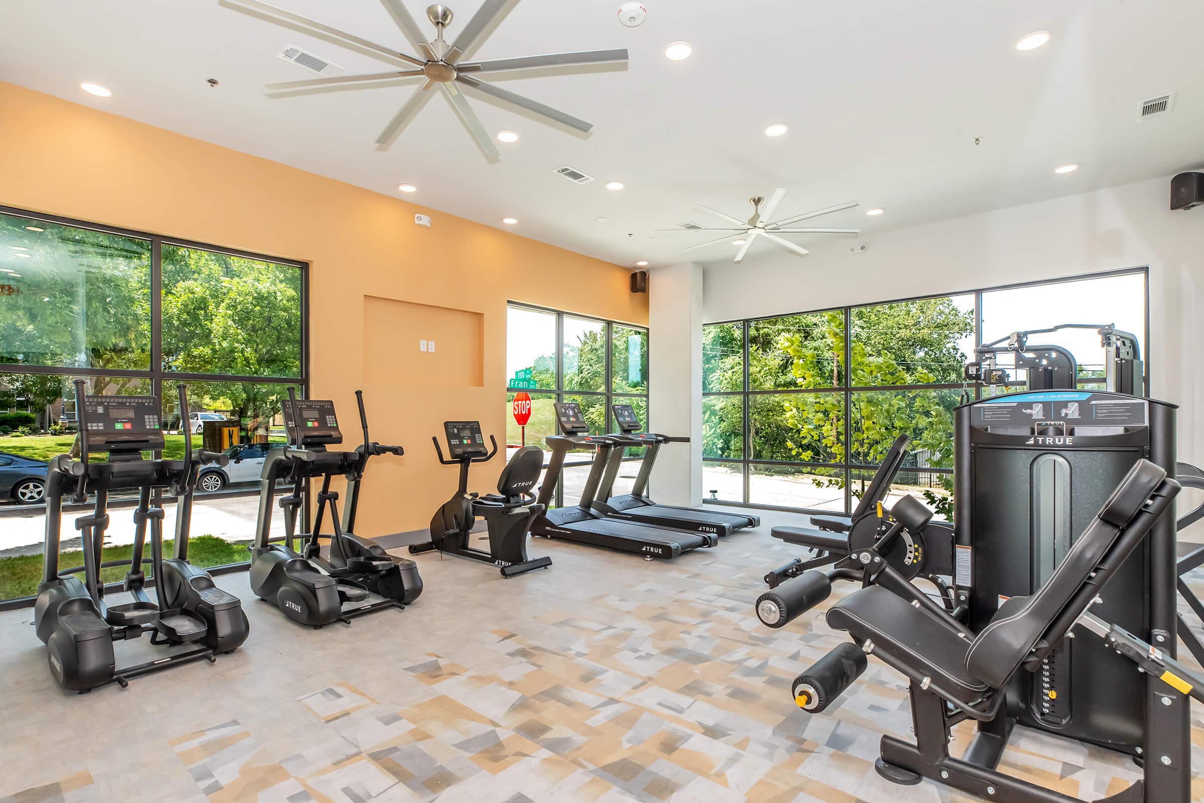 A bright and spacious gym area featuring several treadmills, elliptical machines, and strength training equipment. Large windows provide a view of greenery outside, and ceiling fans are installed for ventilation. The interior has a modern design with neutral-colored walls and a well-maintained floor.