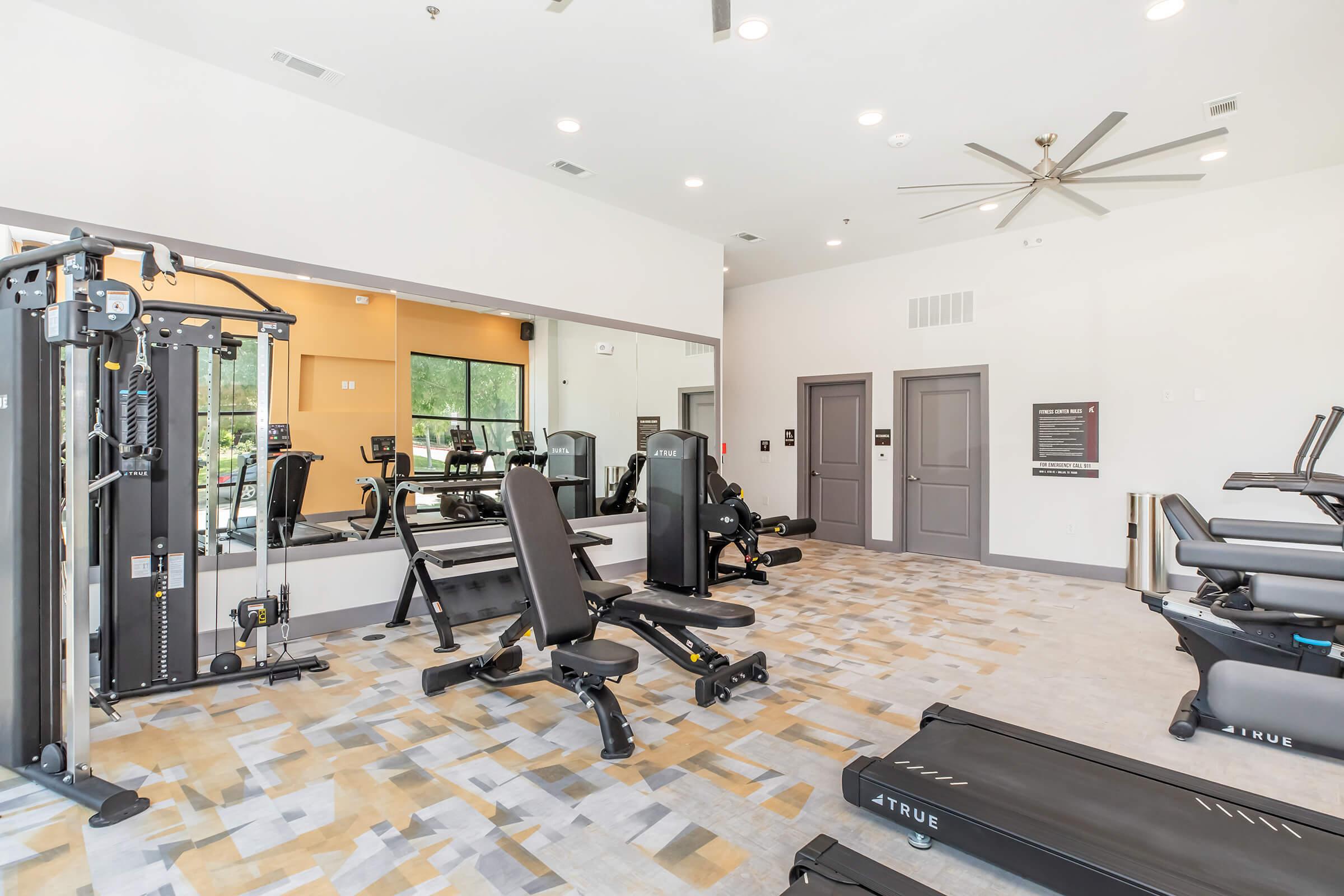 A modern fitness room featuring gym equipment including weight machines, benches, and treadmills. The space is well-lit with large mirrors and a ceiling fan, showcasing a clean, inviting environment for workouts. The flooring has a contemporary design in neutral tones.