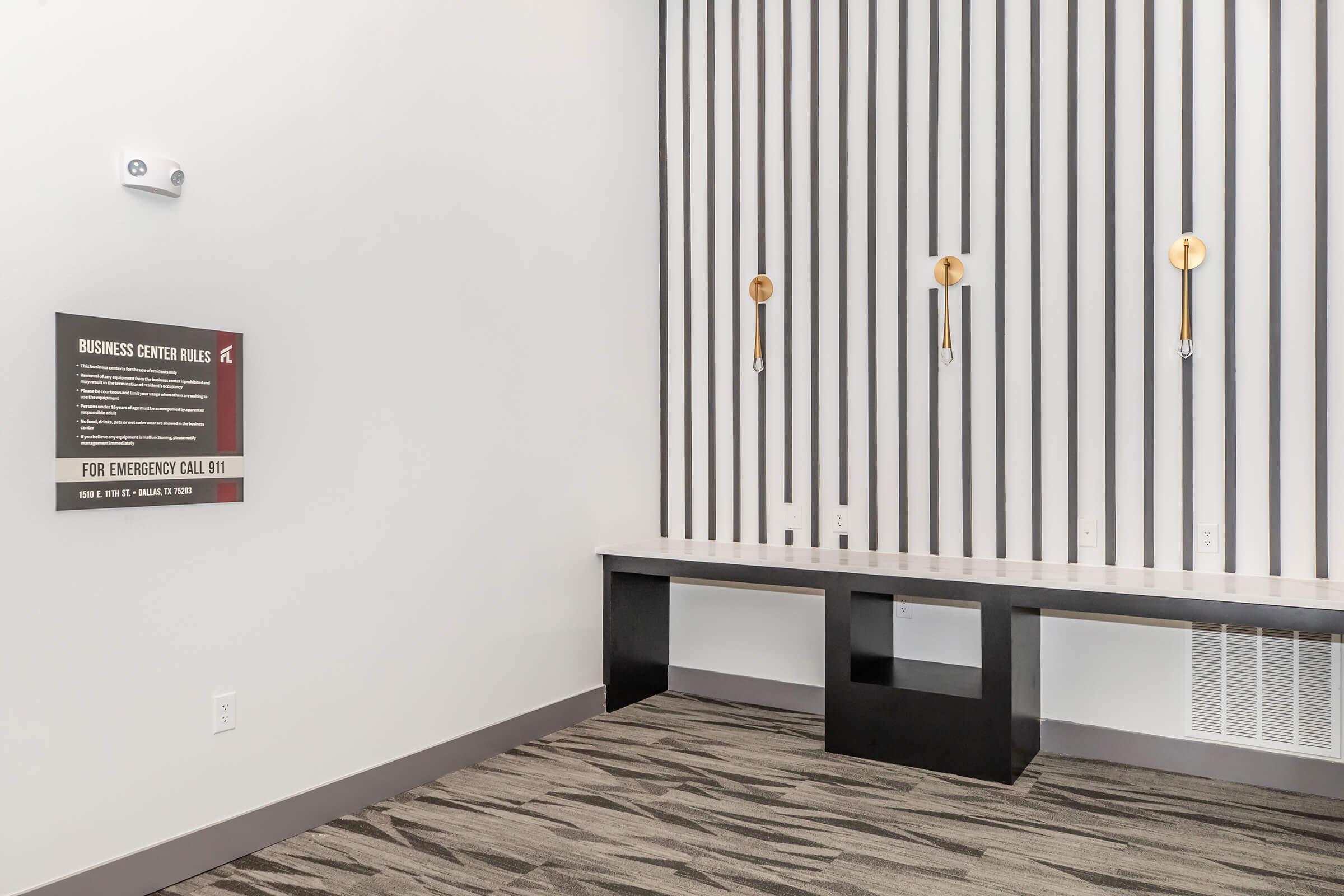 A modern interior of a business center featuring a minimalist design with vertical striped walls. There is a sign listing business center rules on the left and three wall-mounted fixtures above a sleek white counter. The flooring is a textured carpet in neutral tones.