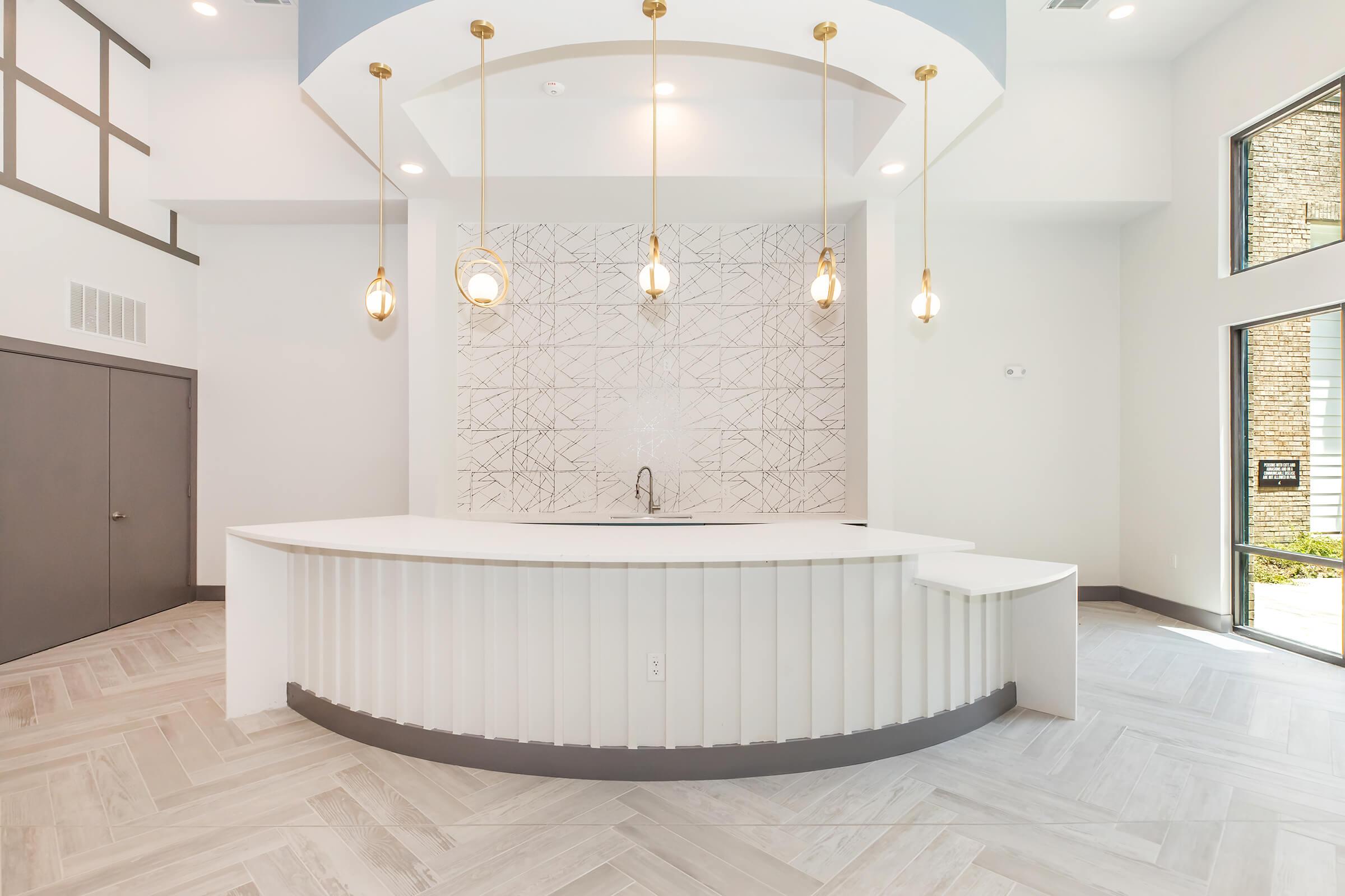 Modern reception area featuring a sleek white curved desk with a geometric patterned backsplash. Three pendant lights hang above the desk, illuminating the space. The floor is composed of light-colored wood planks, and there are large windows allowing natural light to enter, creating a bright and inviting atmosphere.