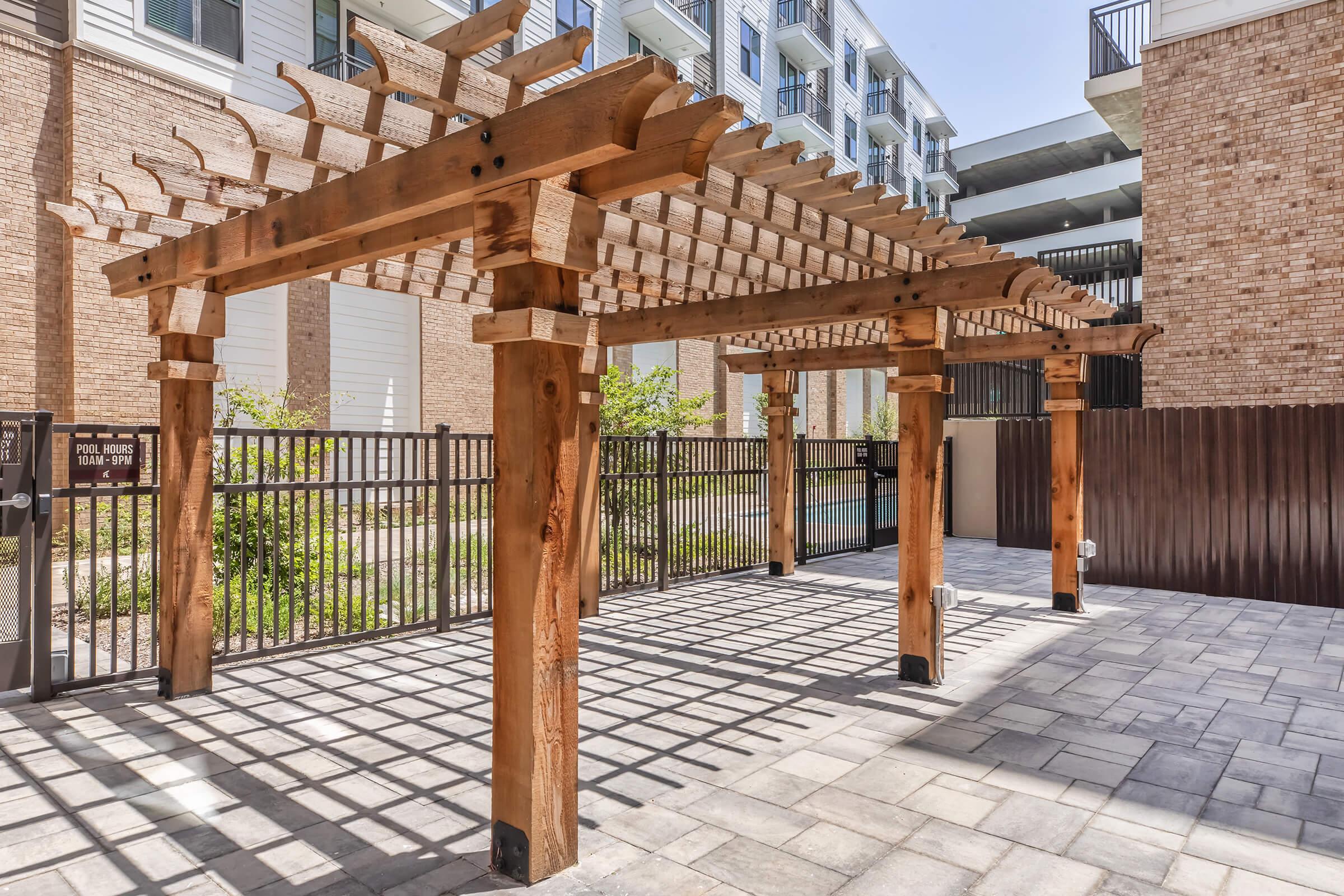 A wooden pergola with a slatted roof stands over a paved area, casting intricate shadows on the ground. In the background, there are modern apartment buildings and landscaping, suggesting a recreational outdoor space. The setting is bright and well-maintained, ideal for relaxation.