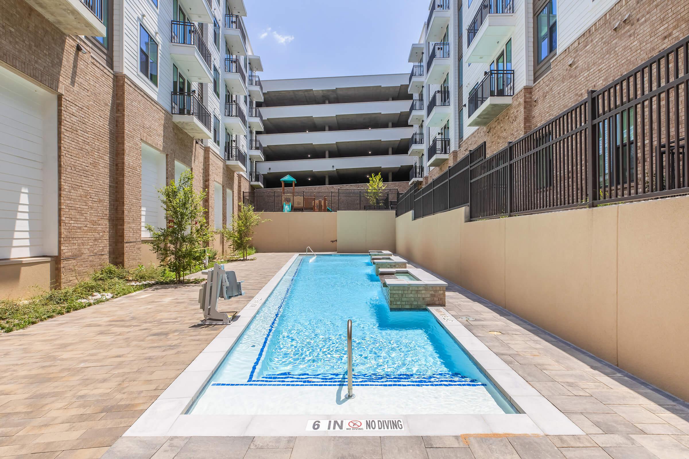 A swimming pool surrounded by modern apartment buildings, featuring a blue water surface and a decorative stone edge. The pool has a "No Diving" sign and is bordered by paved walkways and greenery, creating a relaxing outdoor space.
