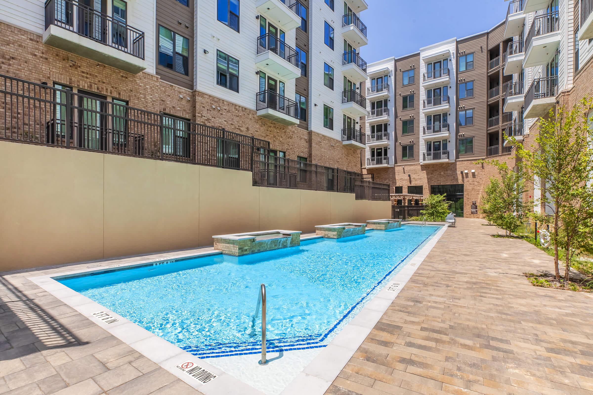 A bright, inviting outdoor swimming pool surrounded by paved decking, with a few small spa areas beside it. The pool is nestled between modern apartment buildings featuring balconies and greenery, creating a serene and attractive living environment.