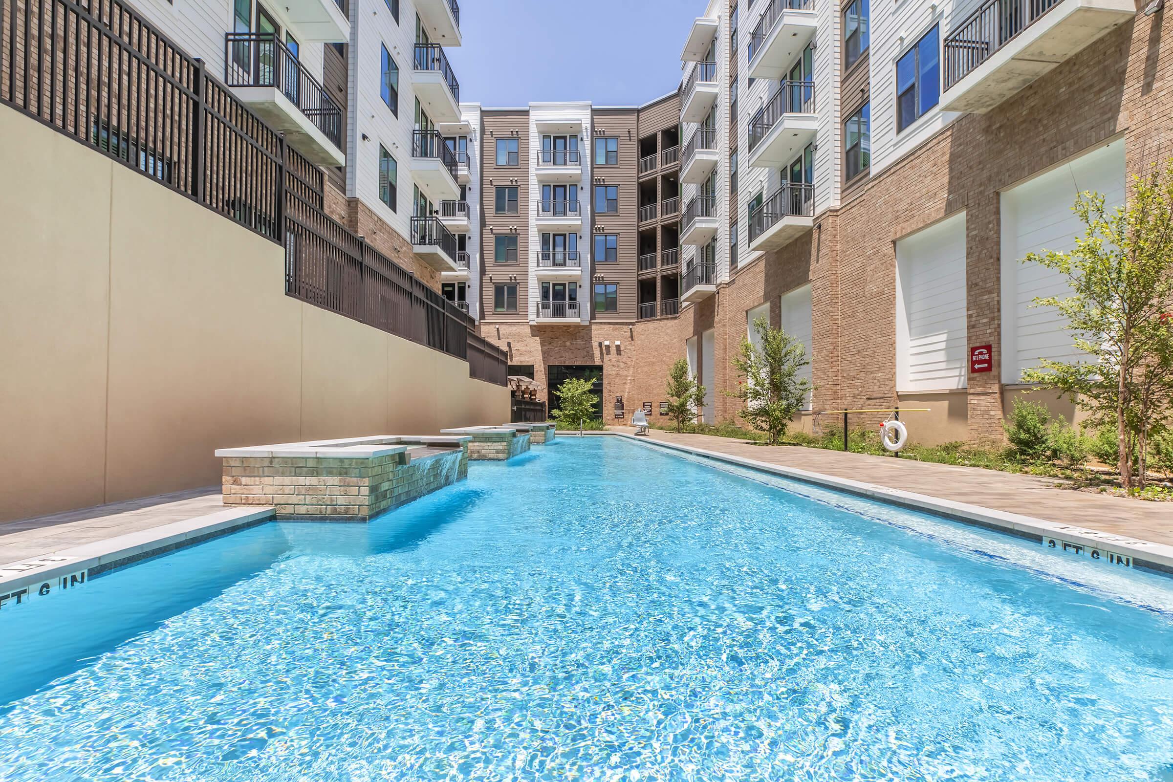 A clear blue swimming pool surrounded by modern apartment buildings. The pool features a pathway alongside, with greenery and trees on either side, creating a serene outdoor space. The sun is shining brightly in a clear sky, highlighting the inviting atmosphere of the area.
