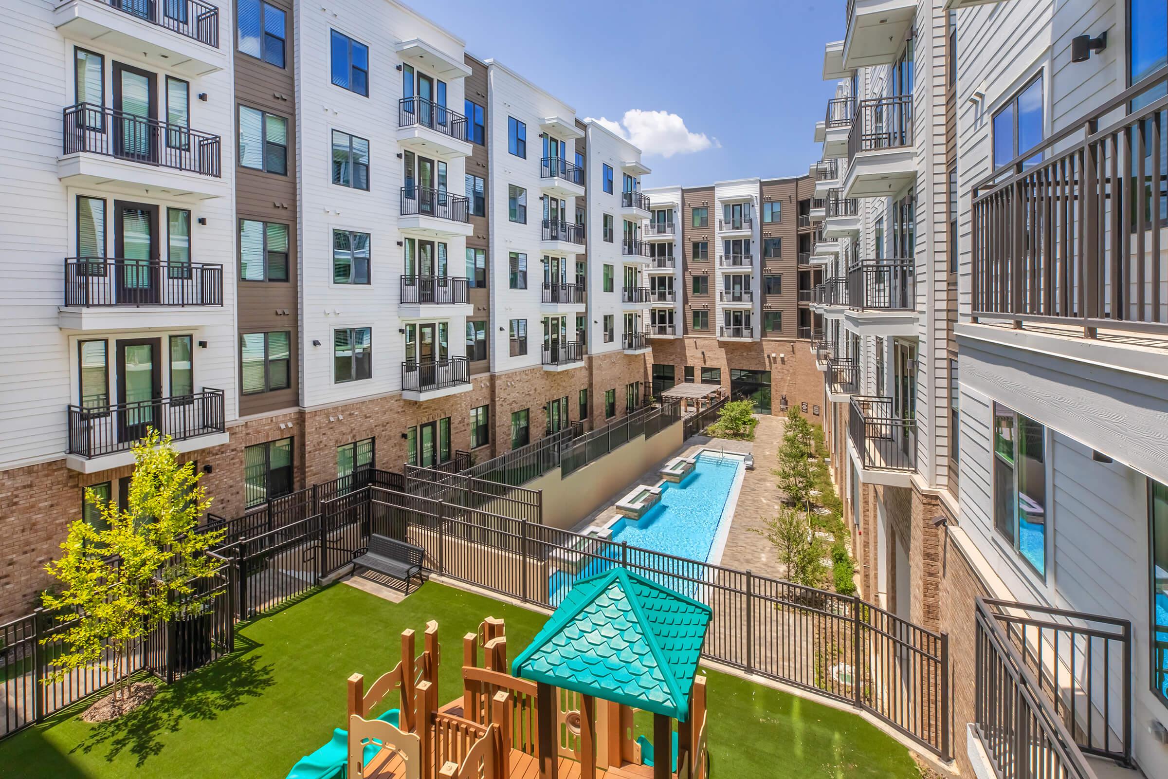 View of a modern apartment complex featuring multiple buildings with balconies. In the foreground, a children's play area with a wooden structure and green turf. A swimming pool runs between the buildings, surrounded by a fence and landscaping, creating a vibrant community space. Clear blue sky overhead.