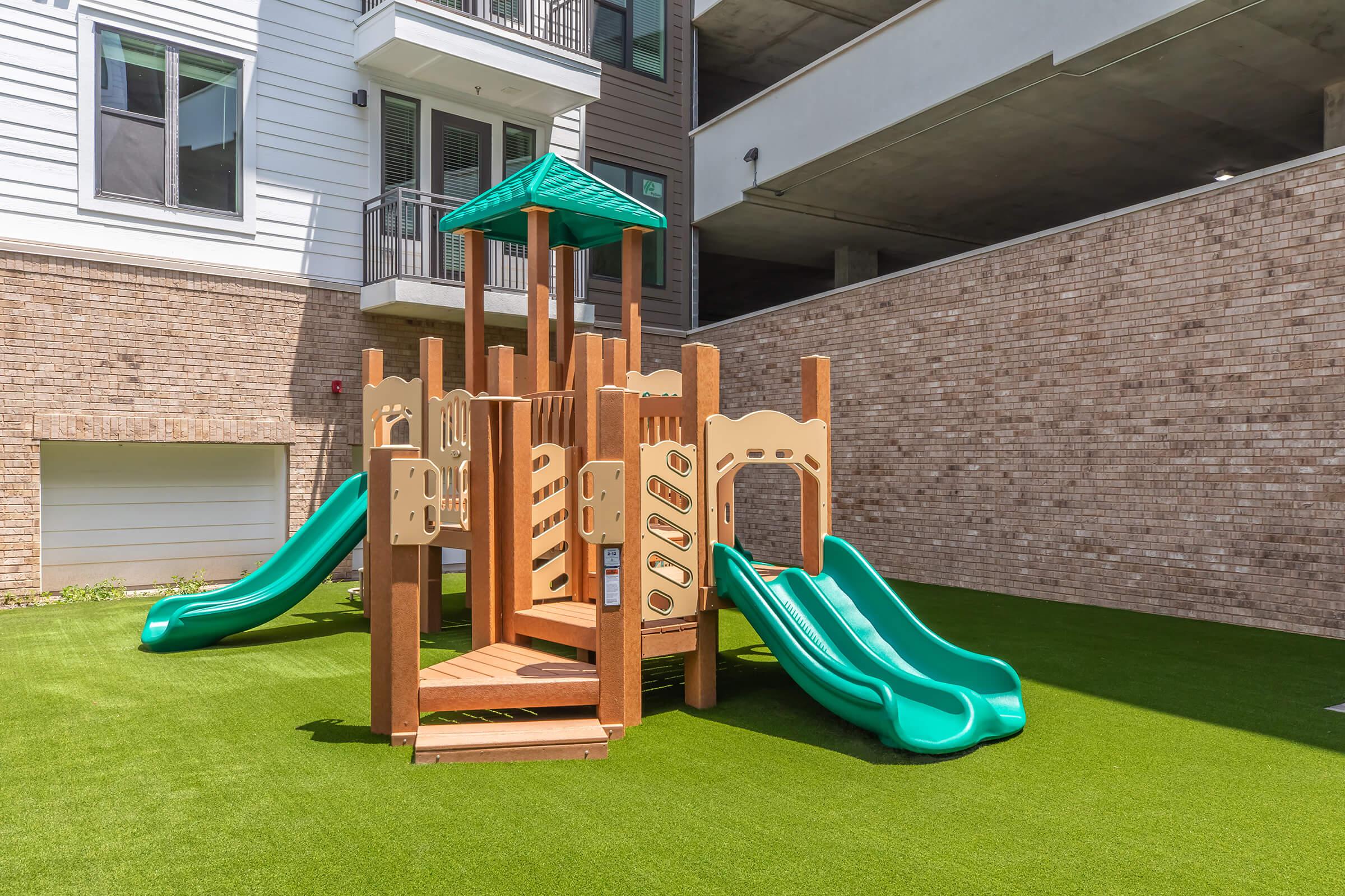 A colorful children's playground structure featuring two green slides, climbing features, and a small roof, situated on artificial grass in a well-lit outdoor area. The background shows a modern building with a parking structure and residential units.