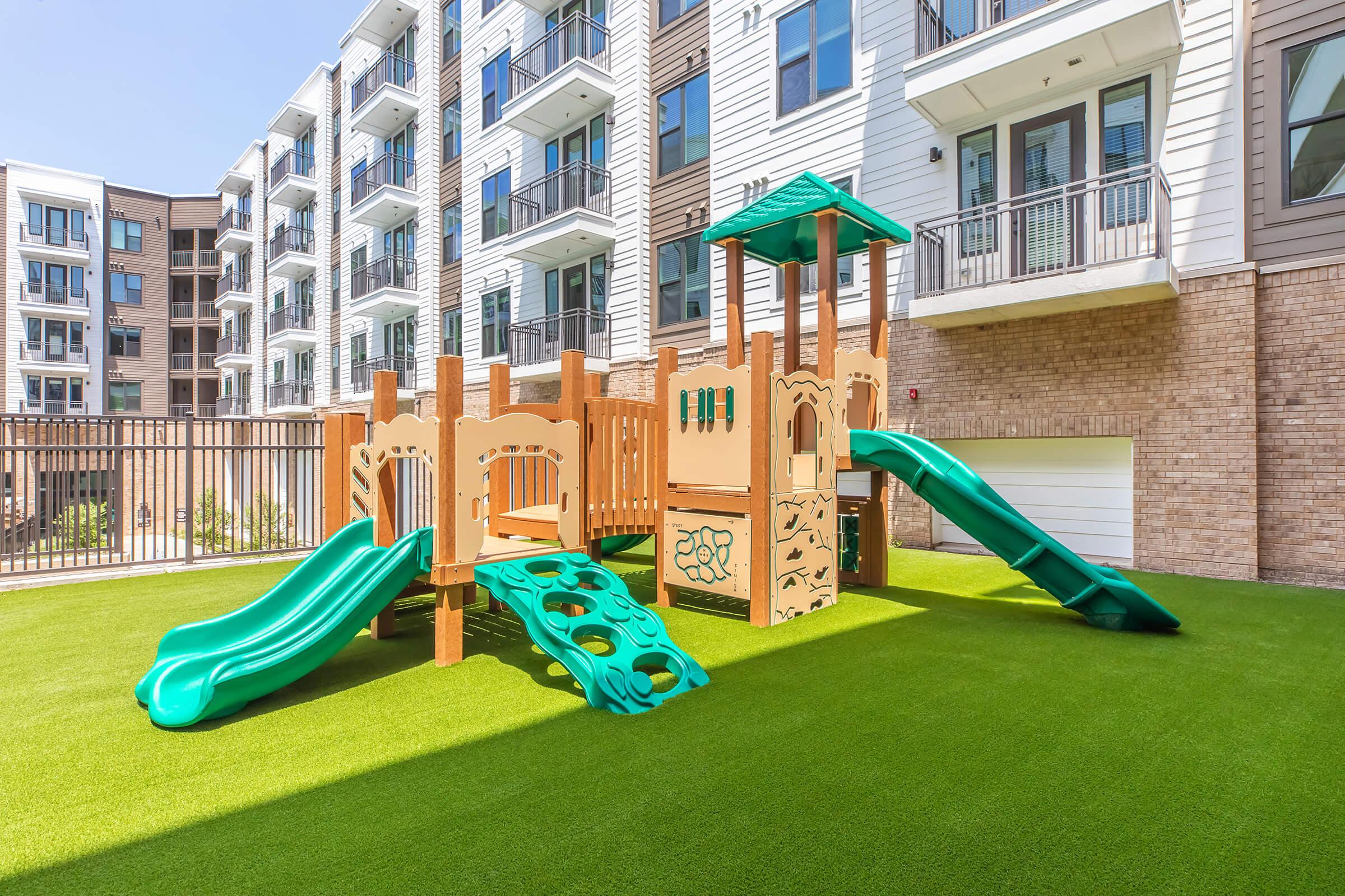 Children's playground with colorful slides and climbing structures, situated on a green artificial turf. The playground is designed for young kids and is located in a courtyard surrounded by modern apartment buildings. Bright and inviting space for outdoor play and recreation.