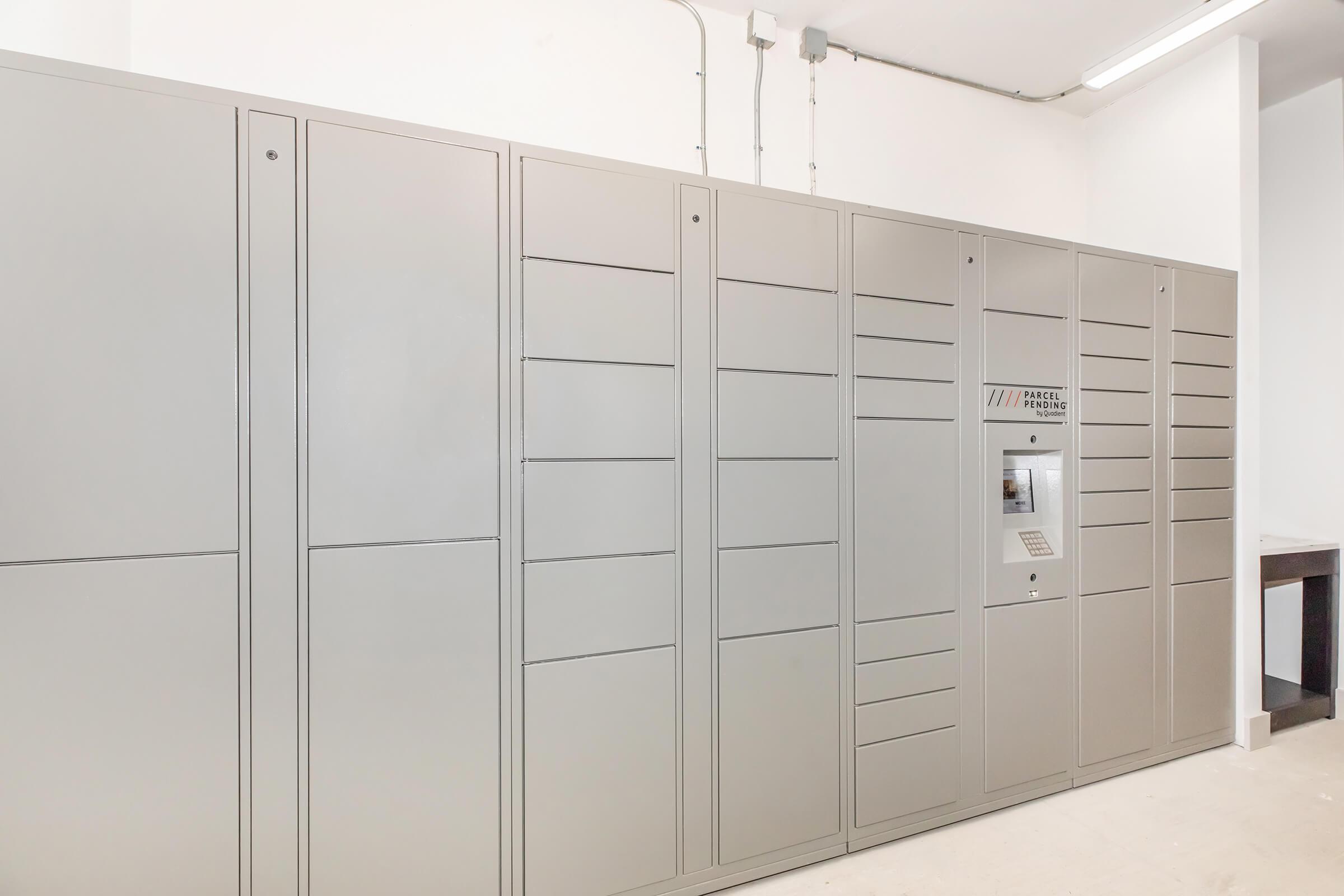 A gray parcel locker system installed in a white-walled room, featuring multiple compartments for package storage. There is a central touch screen panel for user interaction, and the overall setup appears modern and functional. The area is well-lit with minimal furnishings.