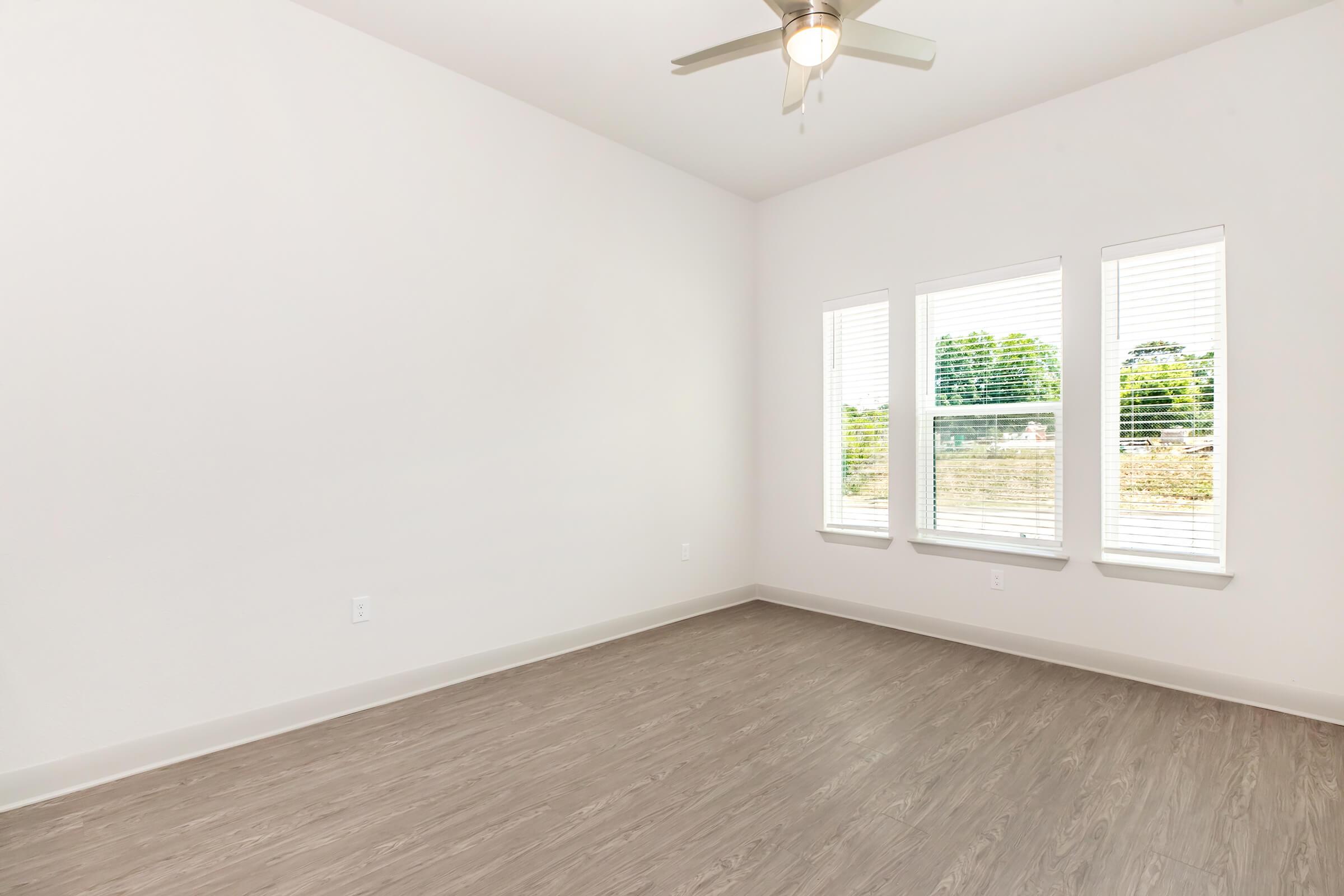 Bright and airy room featuring light-colored walls, a ceiling fan, and large windows with blinds. The flooring is a light wood laminate, and the space is empty, creating an inviting atmosphere.