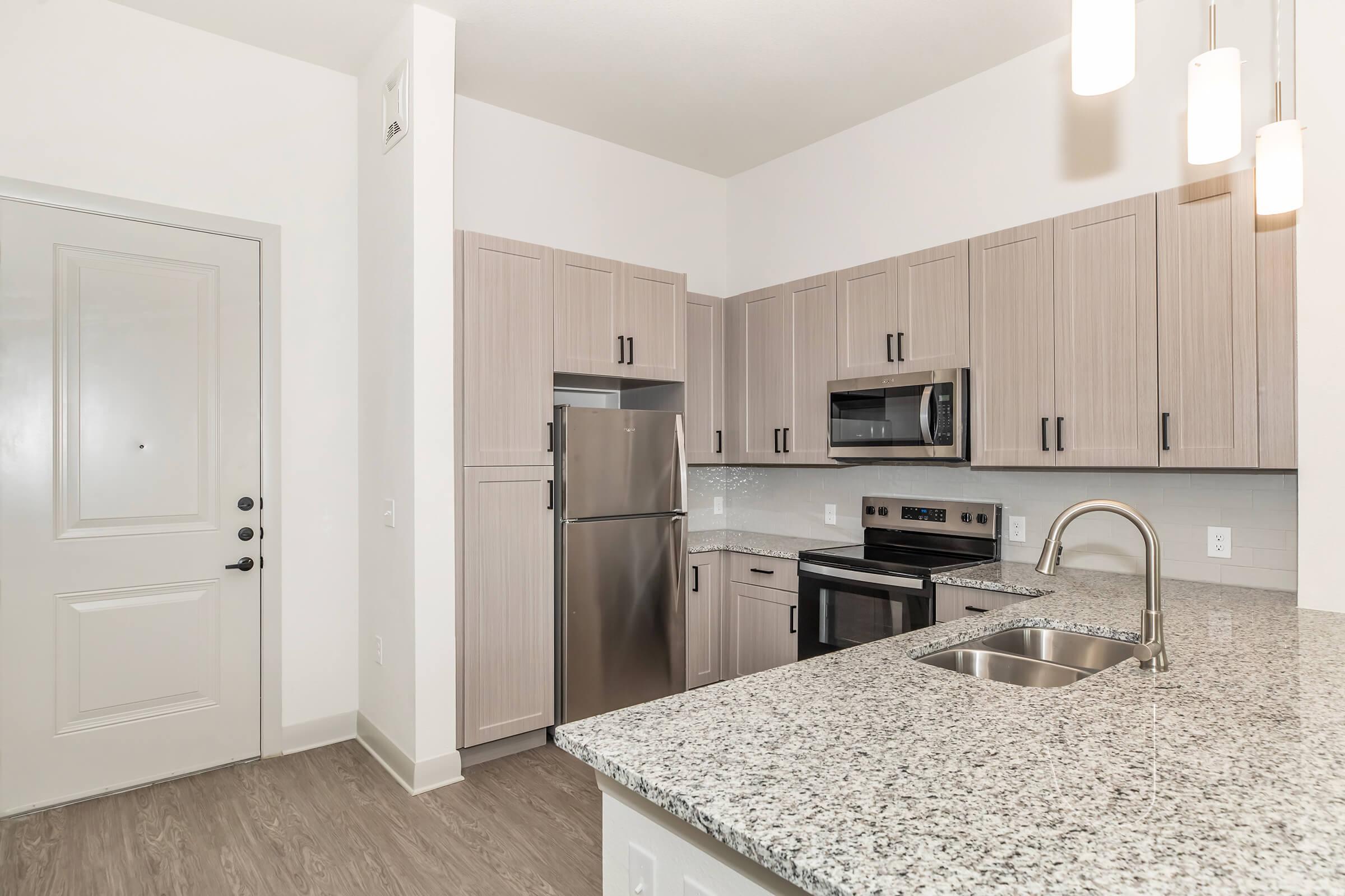 Modern kitchen featuring light wood cabinets, stainless steel appliances including a refrigerator, microwave, and stove, along with a granite countertop and an undermount sink. The space has a clean and contemporary design, with pendant lighting and a neutral color palette. A door is visible in the background.