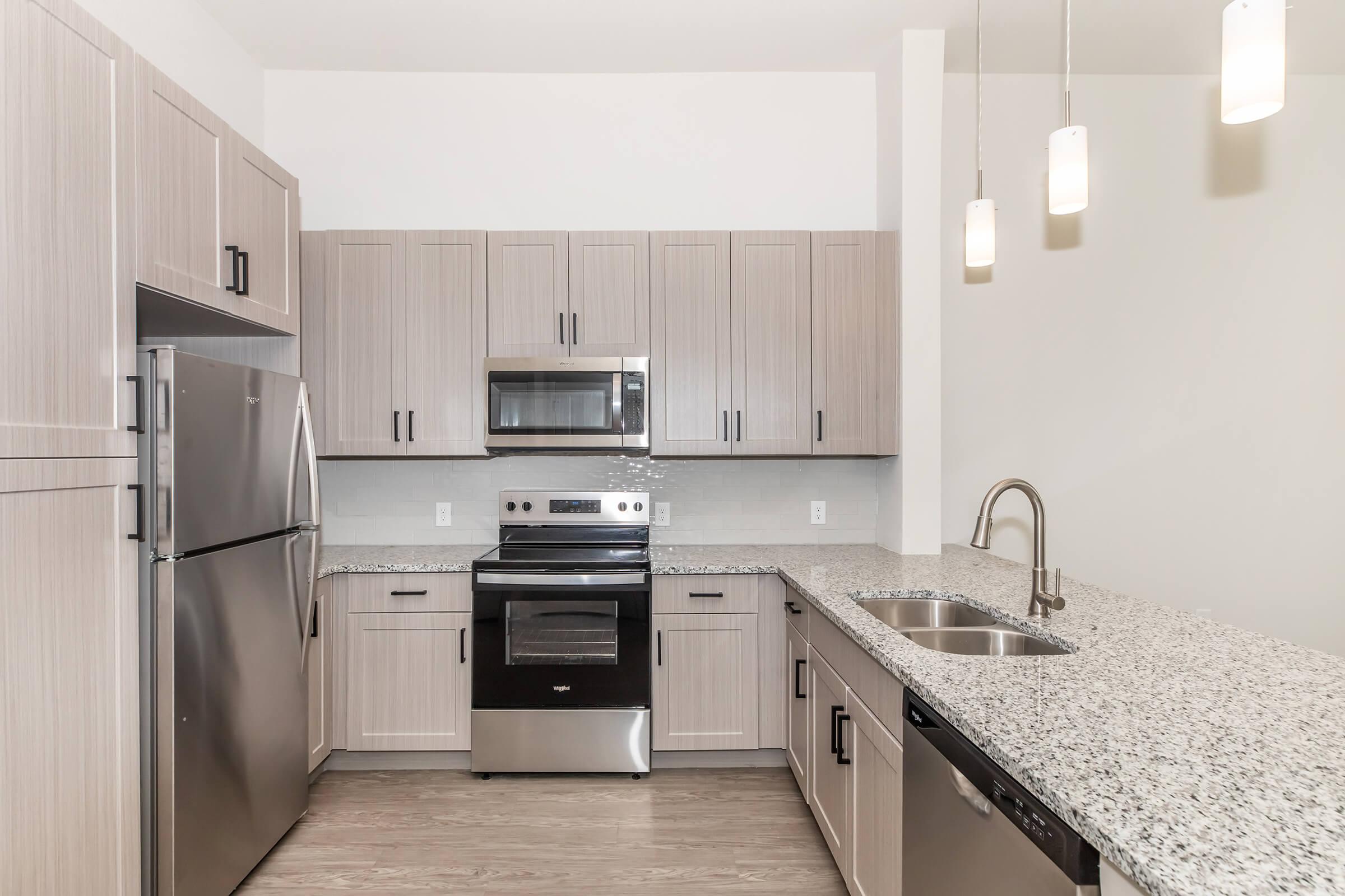 Modern kitchen featuring light wood cabinets, stainless steel appliances including a refrigerator, oven, and microwave, granite countertops, and a double sink. The space is illuminated by pendant lights hanging from the ceiling, with a light-colored wall and hardwood flooring.