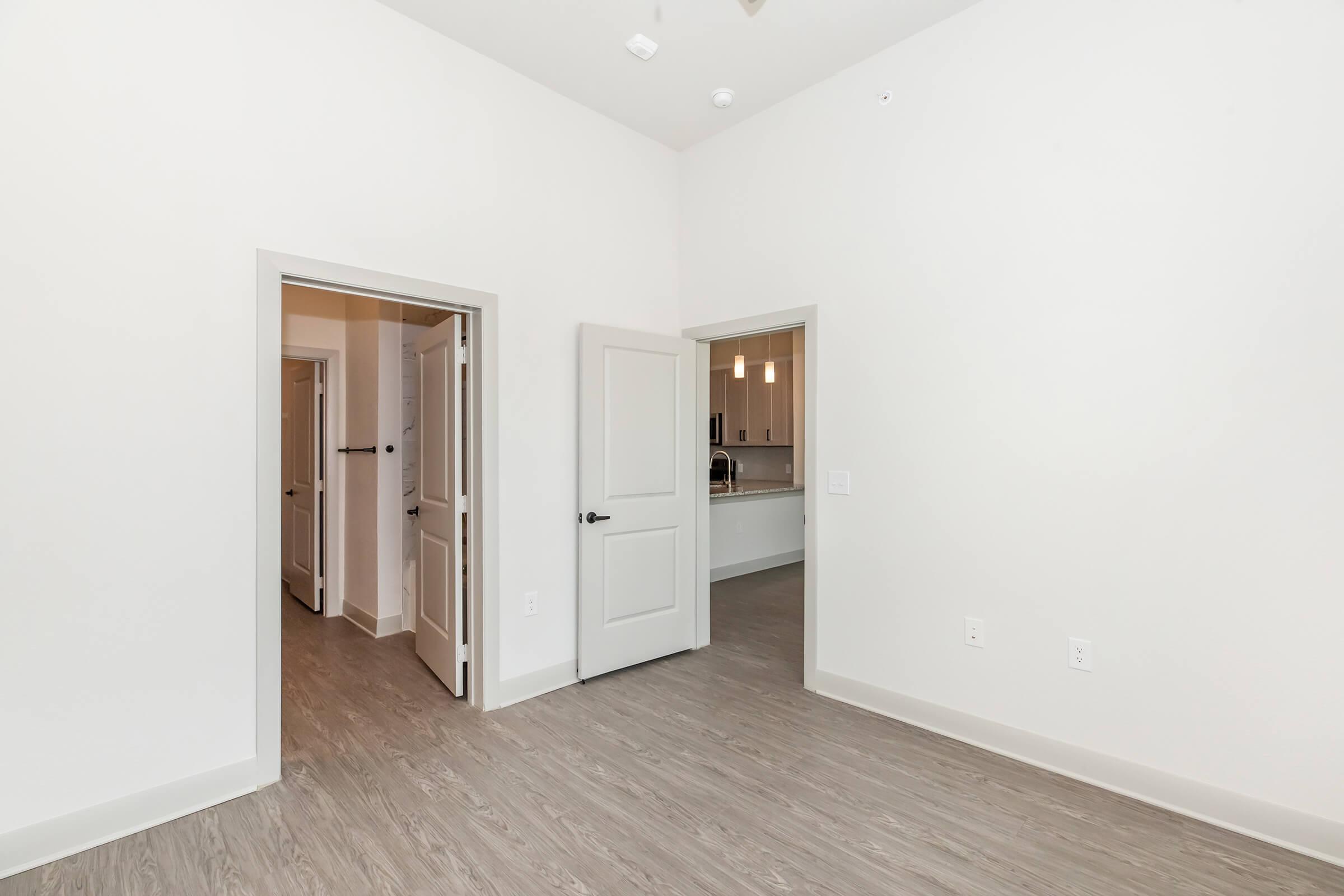 A bright, empty room with light-colored walls and a wooden floor. Two doorways lead to adjacent rooms, suggesting a layout typical of an apartment. The room is minimally furnished, creating a spacious and clean atmosphere.