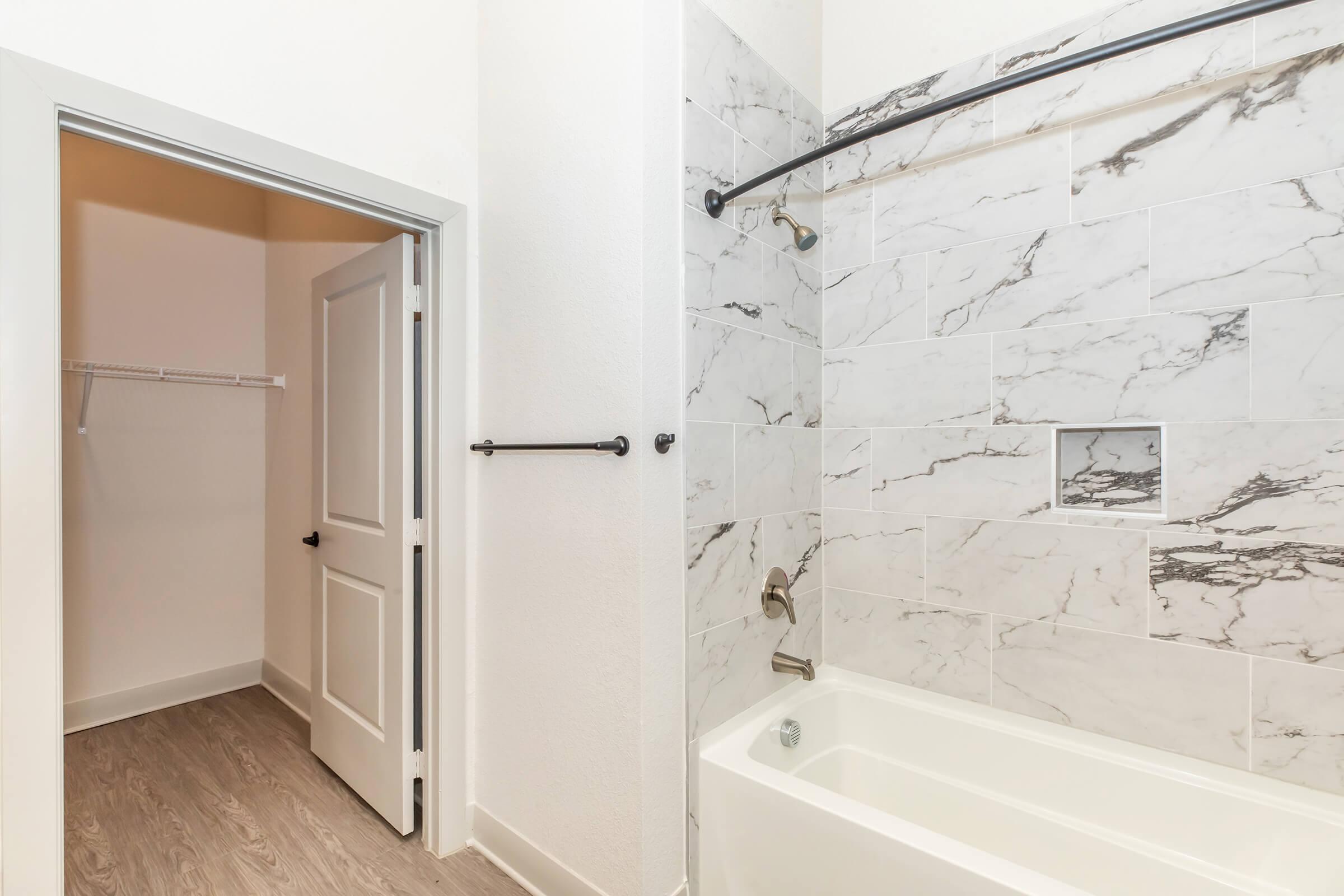 Interior view of a modern bathroom featuring a bathtub and marble wall tiles. A doorway leads to a closet space on the left. The color scheme is light and neutral, with a focus on clean lines and contemporary design elements.