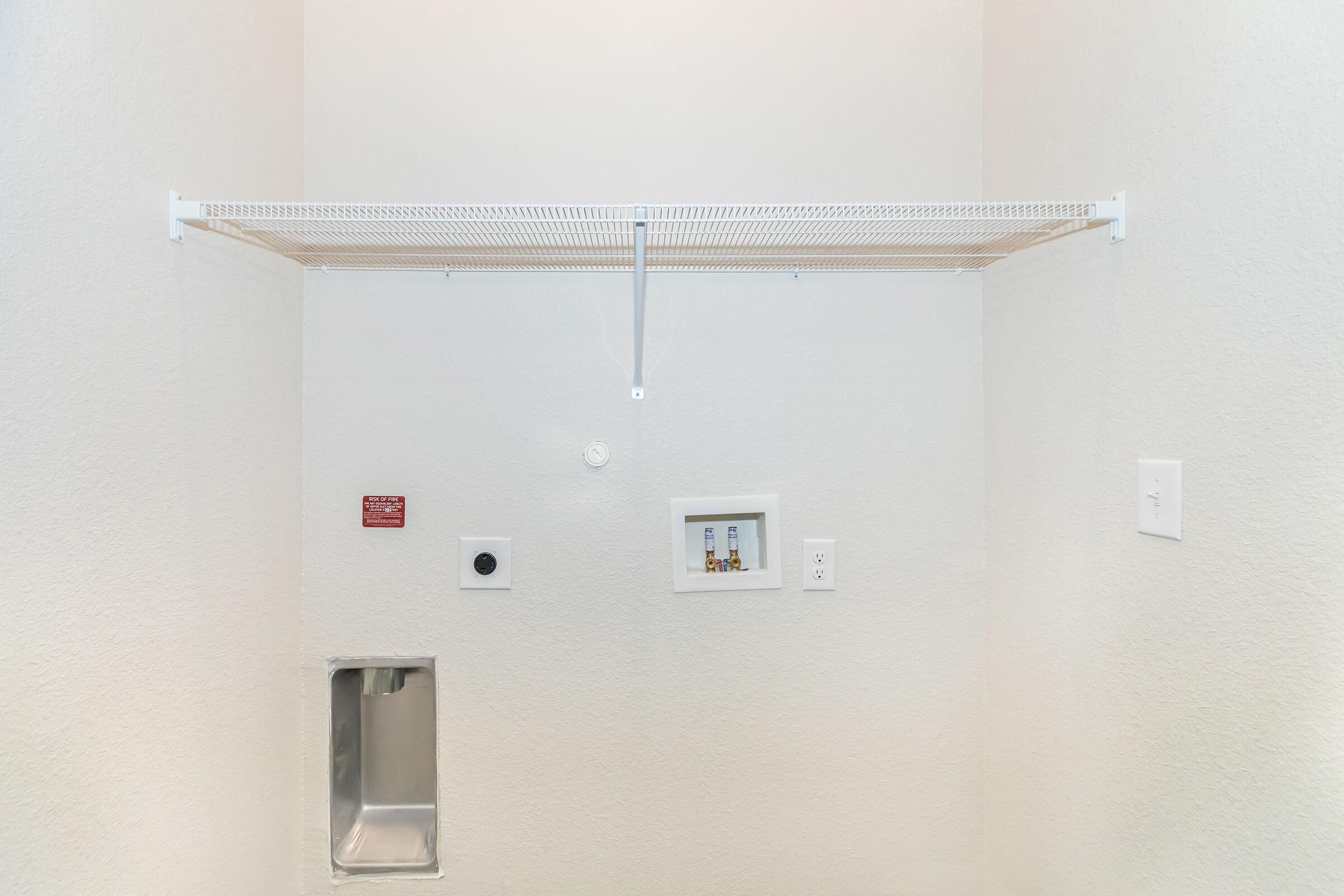 Empty laundry room with a white wall. A metal shelf is mounted on the wall, positioned above a framed picture. There are electrical outlets and a small metal panel on the wall.
