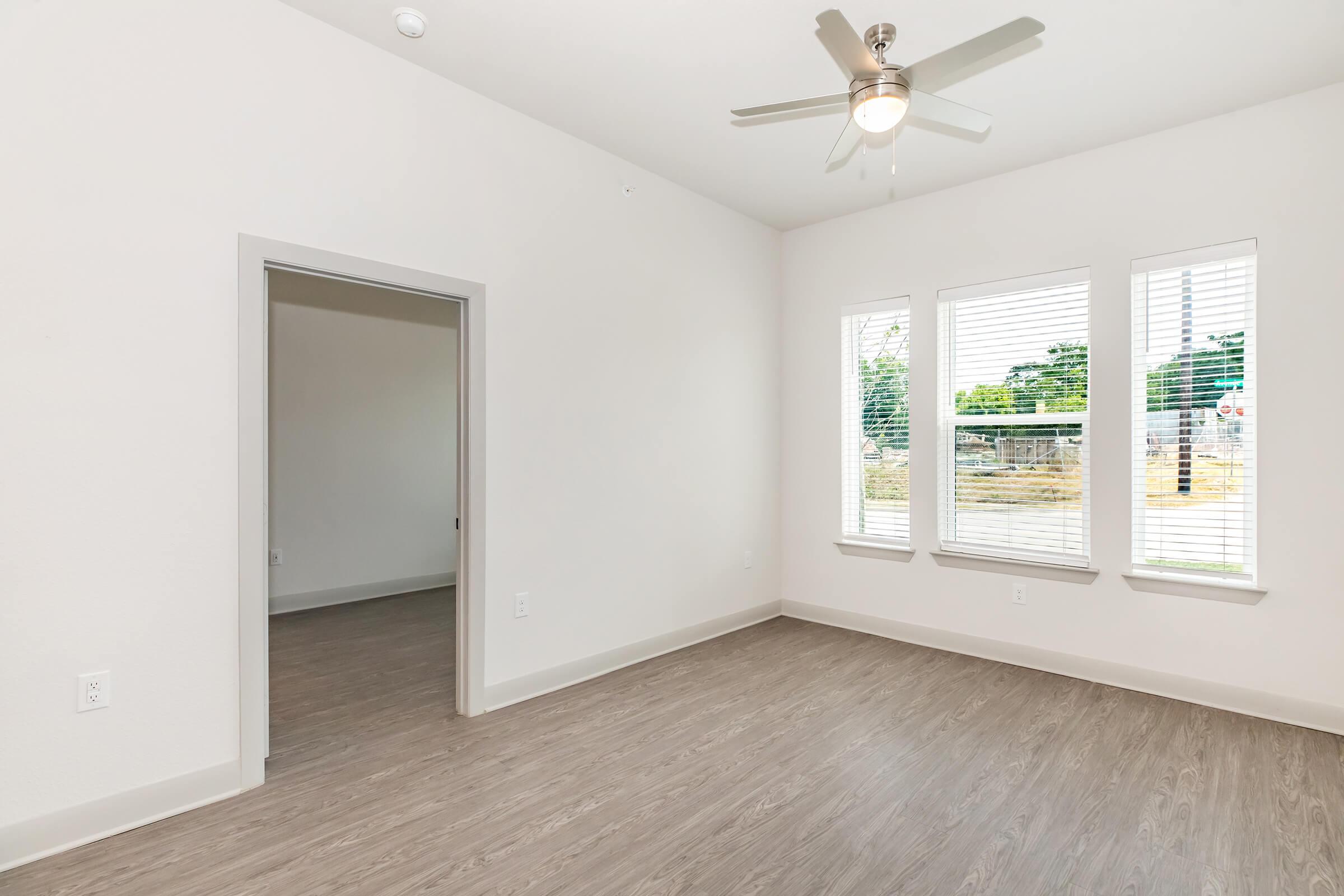 A light and airy room with a ceiling fan, featuring large windows on one side that allow natural light to flow in. The flooring is a light wood finish, and there is an open doorway leading to another space. The walls are painted in neutral tones, creating a bright and welcoming atmosphere.