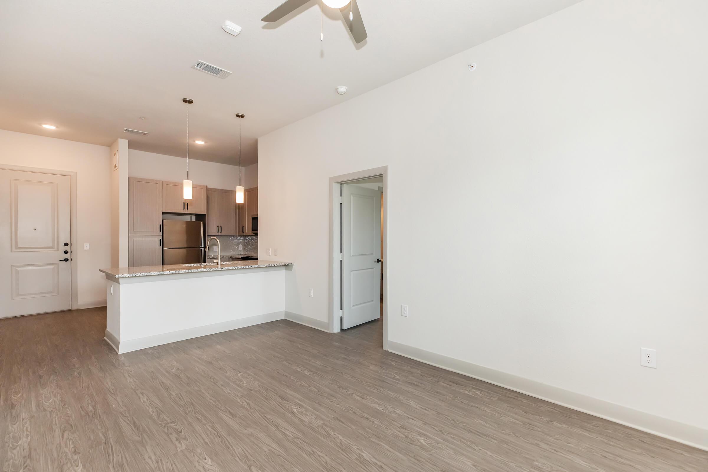 A modern, spacious living area featuring light-colored walls and hardwood flooring. The room includes a ceiling fan and a doorway leading to a kitchen with stainless steel appliances, a granite countertop, and pendant lighting. Natural light fills the space, creating a bright and inviting atmosphere.