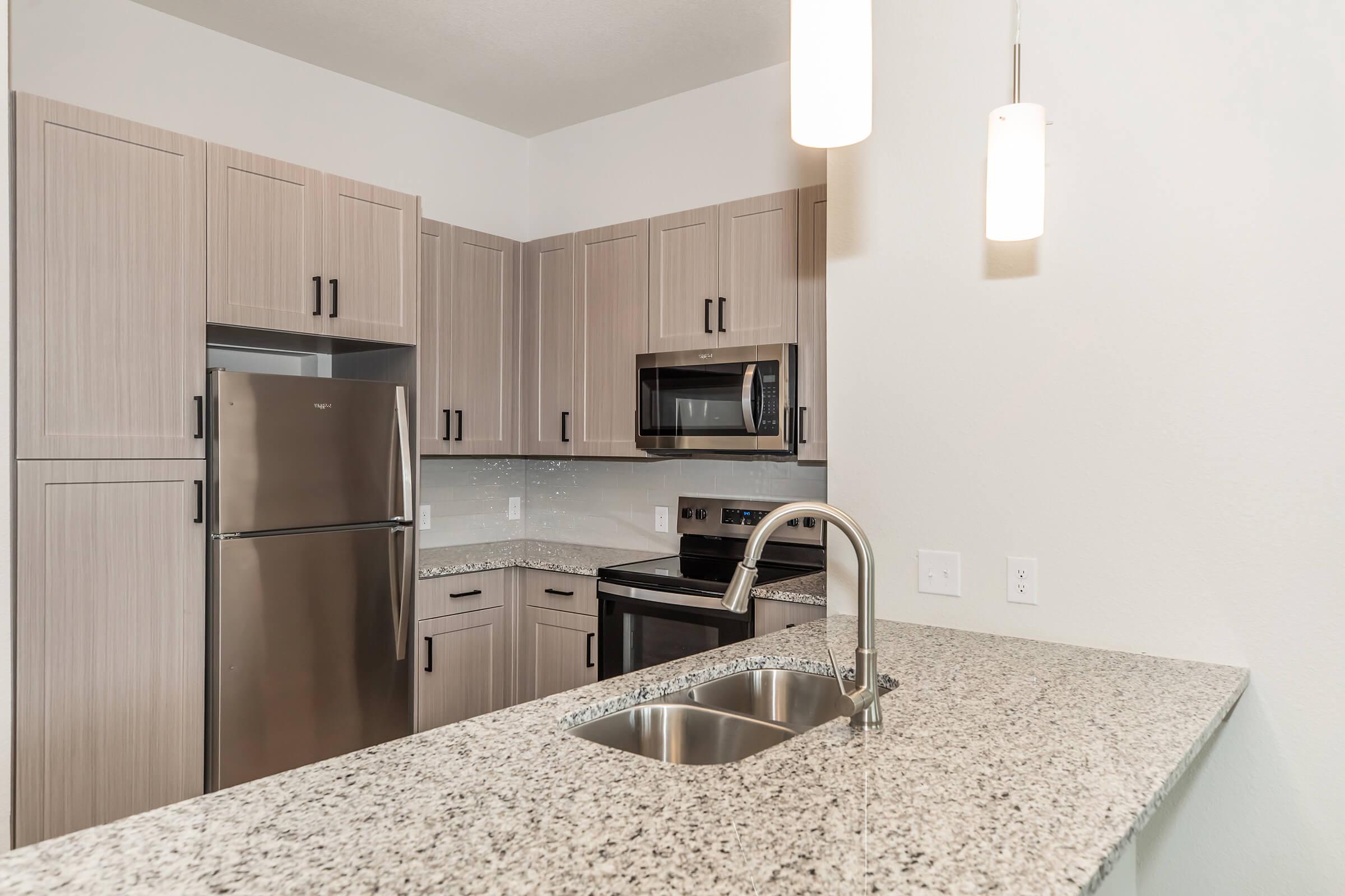 Modern kitchen with light wood cabinetry, stainless steel appliances including a refrigerator and microwave, and a granite countertop. A sink with a sleek faucet is visible, along with pendant lighting hanging above the counter. The overall design is clean and contemporary.