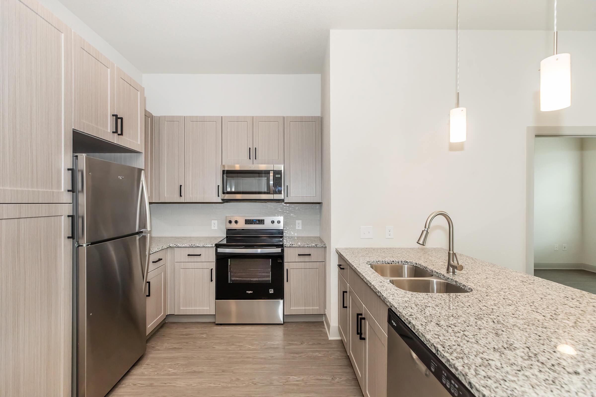 Modern kitchen featuring light wood cabinetry, stainless steel appliances including a refrigerator, stove, and microwave. The countertop is made of gray speckled granite, and there are pendant lights hanging above the sink. The open layout connects to a living area with neutral-colored walls and flooring.