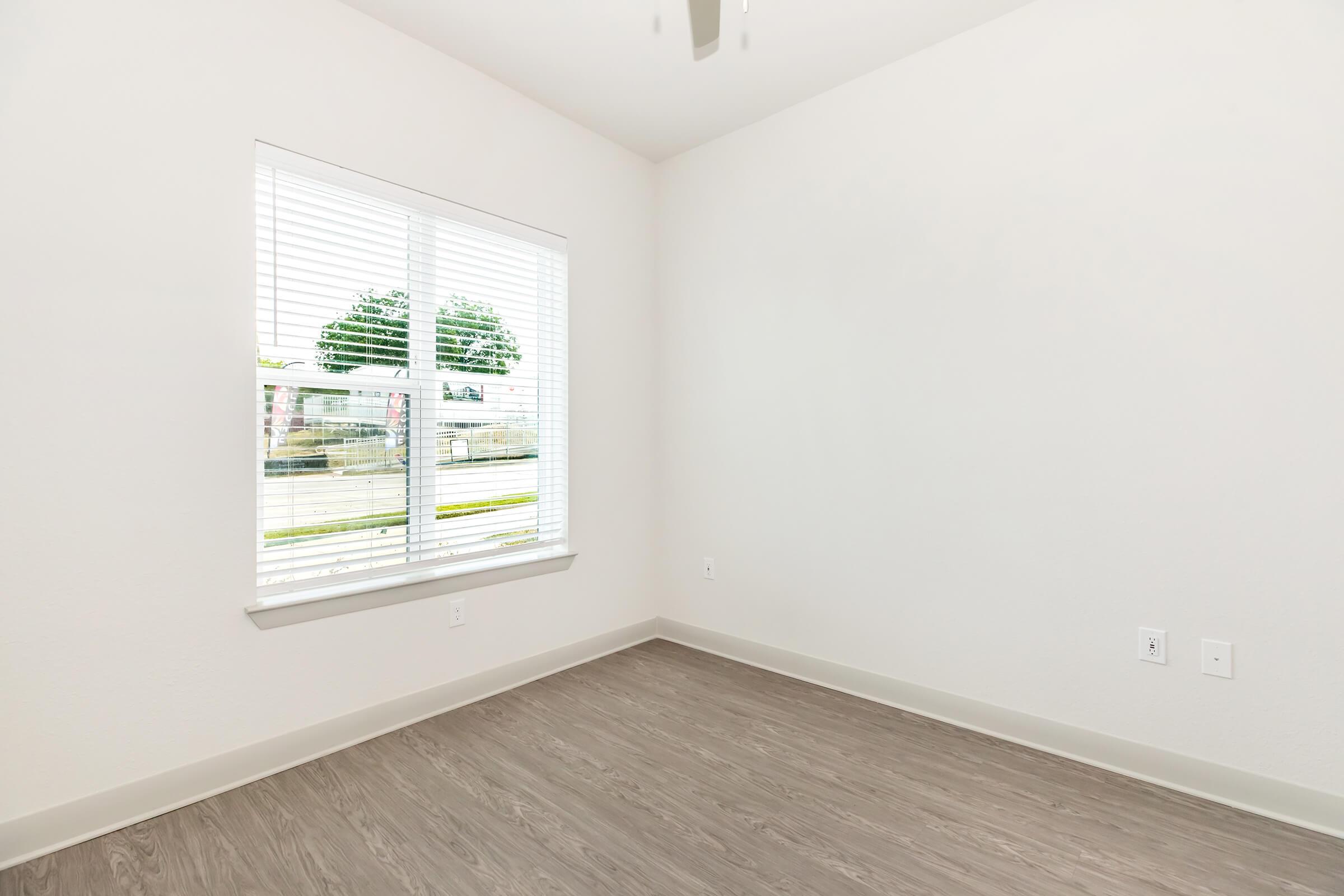 Empty room with light-colored walls and vinyl flooring. A window with blinds allows natural light to enter, showing a view of trees and greenery outside. The space appears clean and minimalistic, suitable for various purposes such as a bedroom or office.