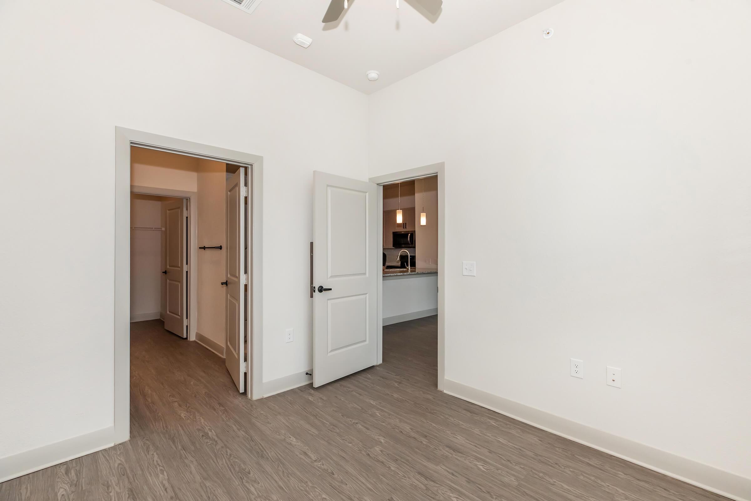 Interior view of a modern room featuring light-colored walls, a wooden floor, and two open doorways leading to adjacent spaces. One doorway opens to a kitchen area, while the other leads to a hallway. Natural light from unseen windows enhances the simplicity of the design.