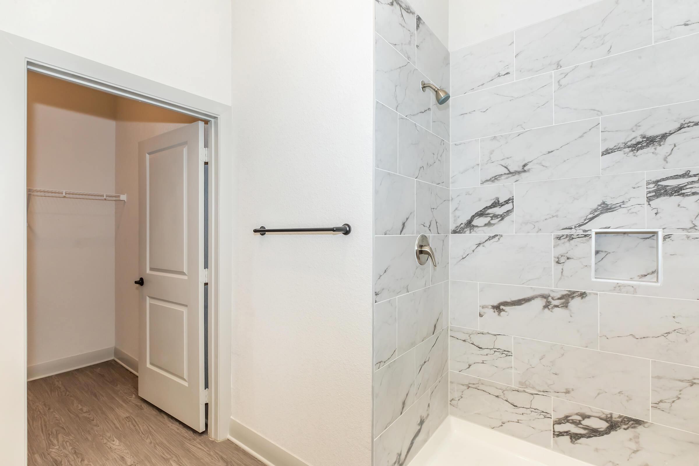 A modern bathroom featuring a walk-in shower with marble tiles, a towel rack on the wall, and an open closet space leading to the room. The space is well-lit and has a clean, contemporary design with light-colored walls and flooring.