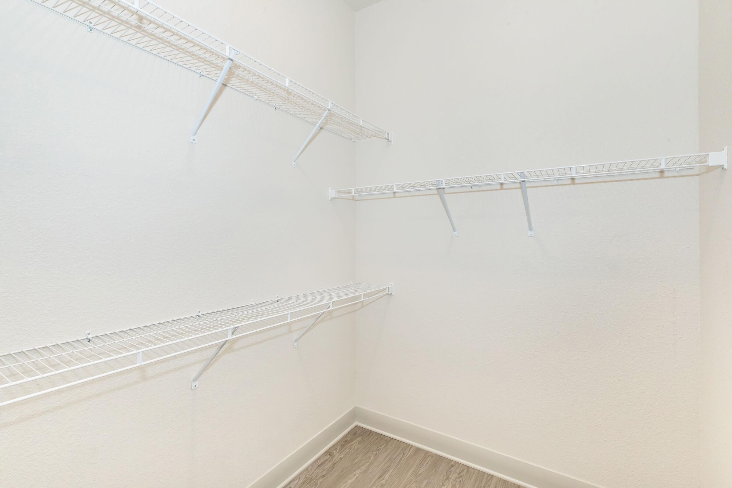A clean, empty closet featuring several white wire shelves mounted on the walls. The walls are painted in a light color, and the floor has a light wood finish, creating a spacious and minimalist look. The shelves provide ample storage options for organizing items.