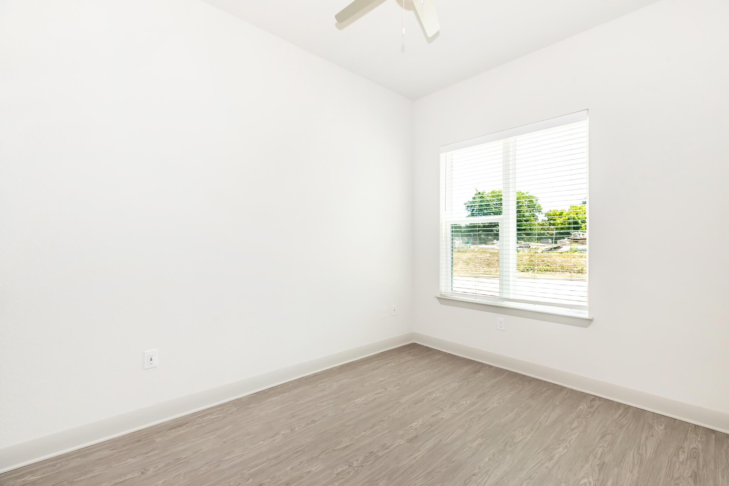 Empty room with light-colored walls and a wooden floor. A window with white blinds lets in natural light, revealing greenery outside. No furniture is present, creating a spacious and minimalist atmosphere. A ceiling fan is visible in the corner.