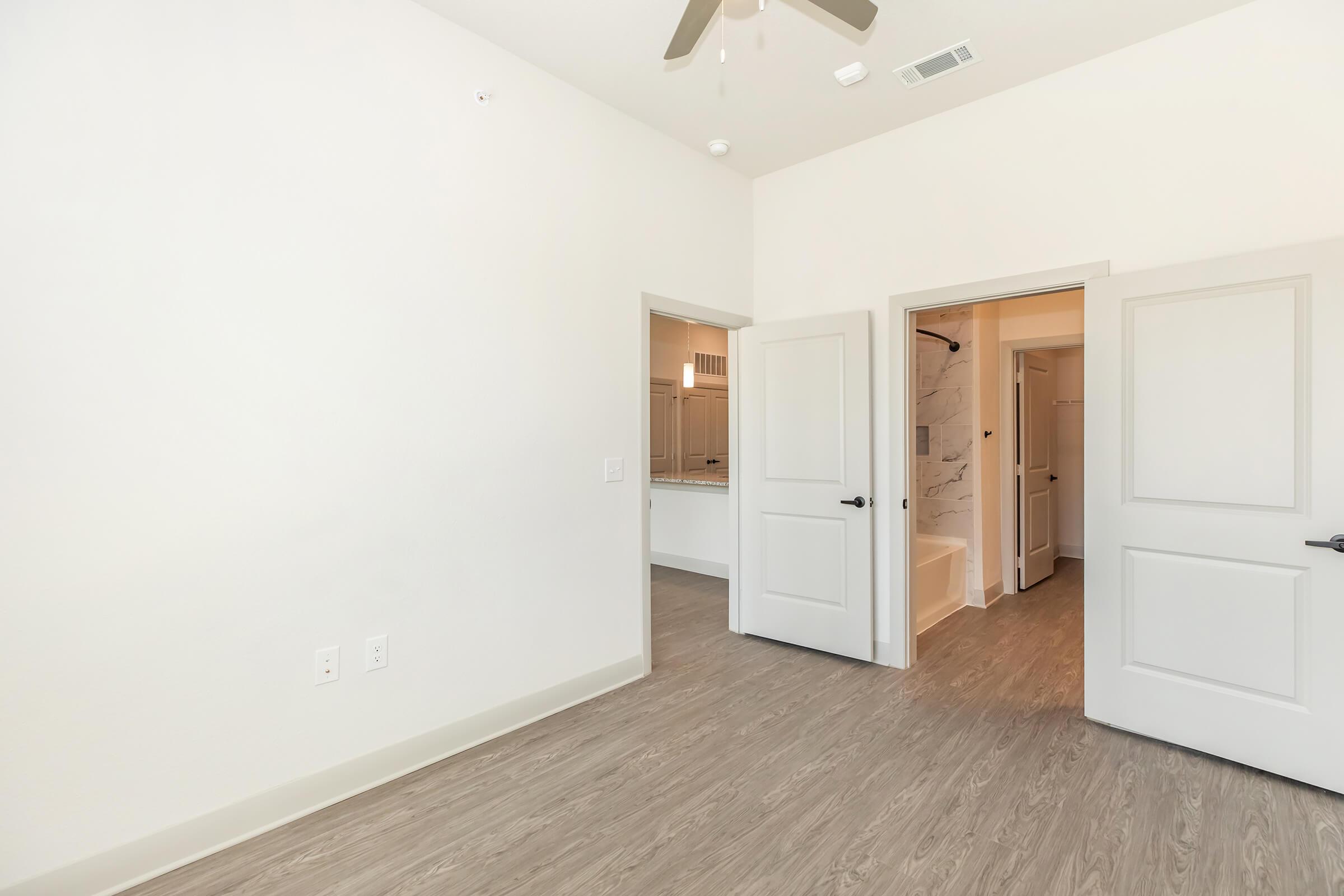 A spacious interior of a room with light-colored walls and a wooden floor. Two open doors lead to adjacent areas, one showing a bathroom with a bathtub and the other possibly leading to a closet or additional space. The ceiling features a ceiling fan, and the overall atmosphere is bright and modern.