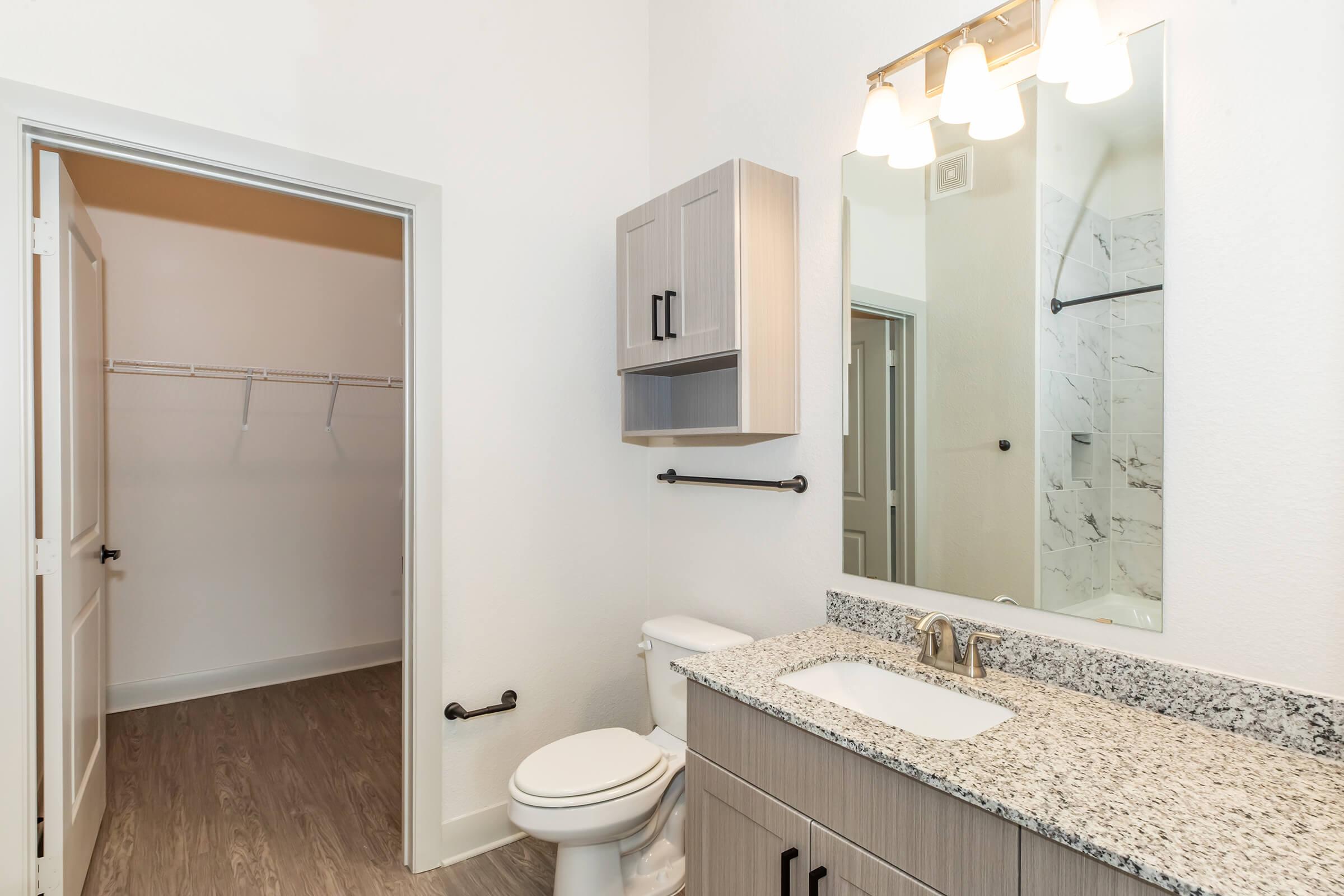 A modern bathroom featuring a granite countertop with a sink, a toilet, and a mirror above the sink. There is a light fixture with three bulbs. A closet with shelves is visible through a doorway on the left. The walls are painted white, and the floor has a light wooden finish.