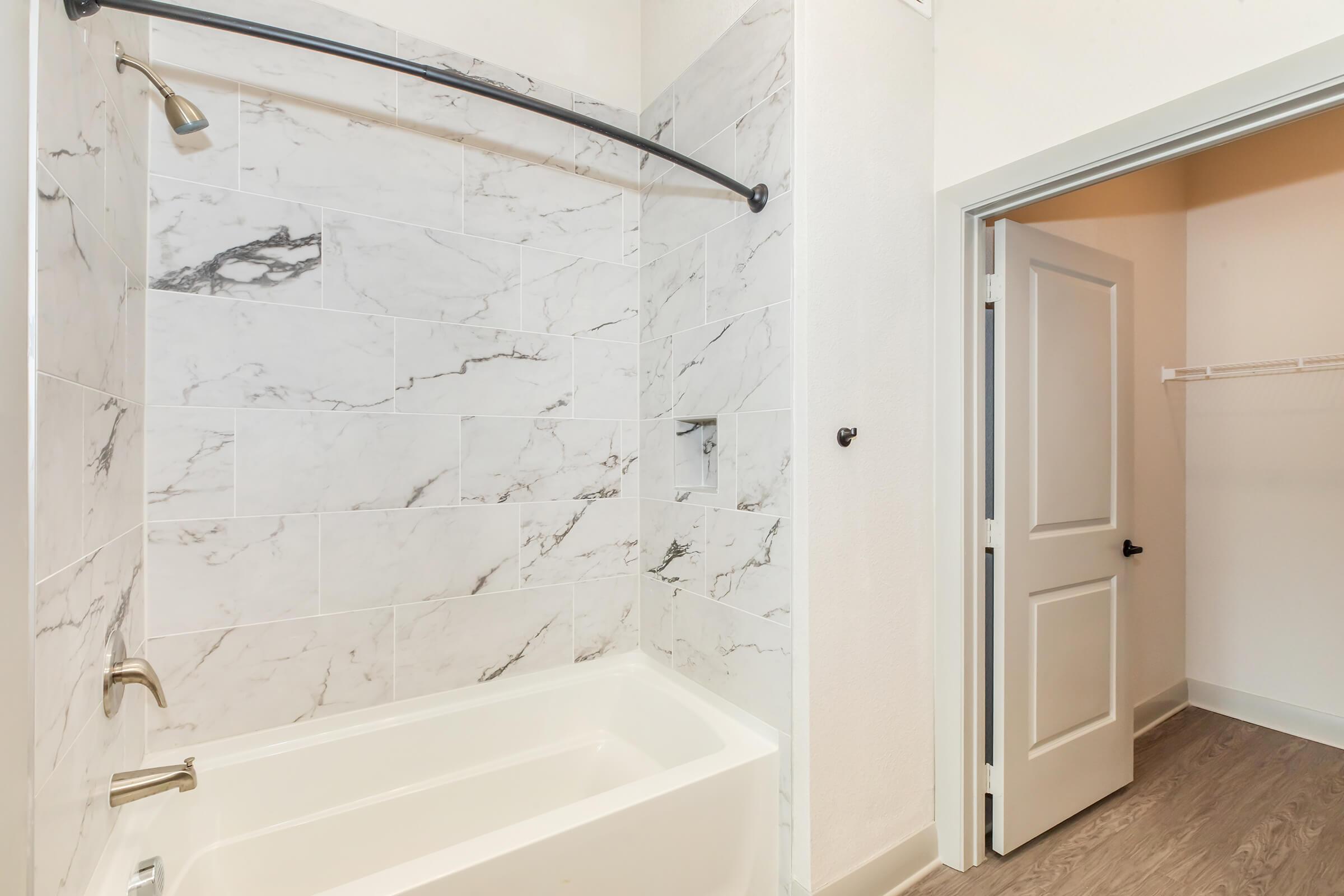 A modern bathroom featuring a white bathtub with a marble-tiled shower area. The walls are decorated with large white tiles with gray veining. A sleek showerhead is mounted on the wall, and there is a door leading to another room in the background. The flooring is a light wood laminate.