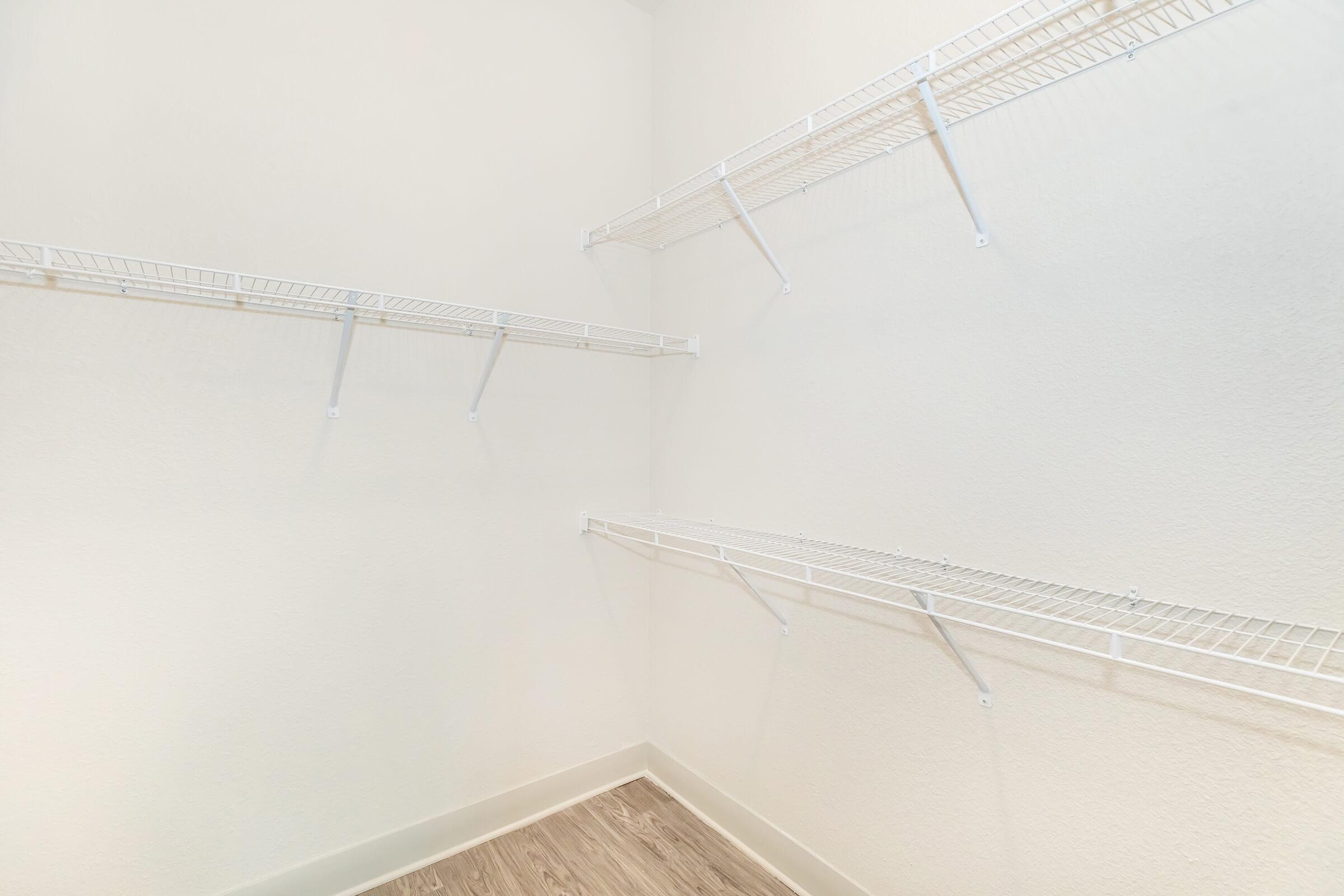 A clean, empty storage closet with white wire shelves mounted on the walls. The walls are painted a light color, and the floor is a light wood or laminate finish. The space is well-lit, providing a bright and airy feel.