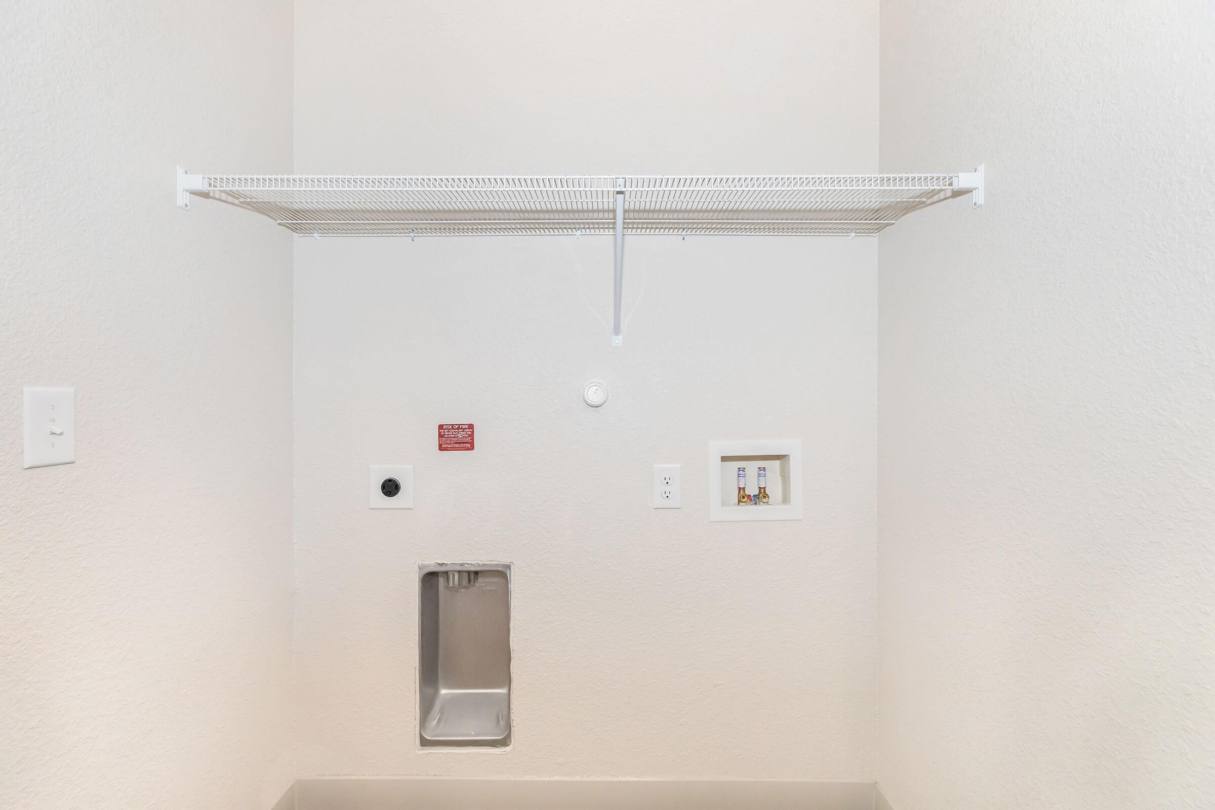 Empty laundry room with a white wire shelf against a light-colored wall. There's a small electrical outlet and a vent on the wall. A metal panel is mounted lower on the wall, and a small, labeled switch or control panel is visible near the floor.
