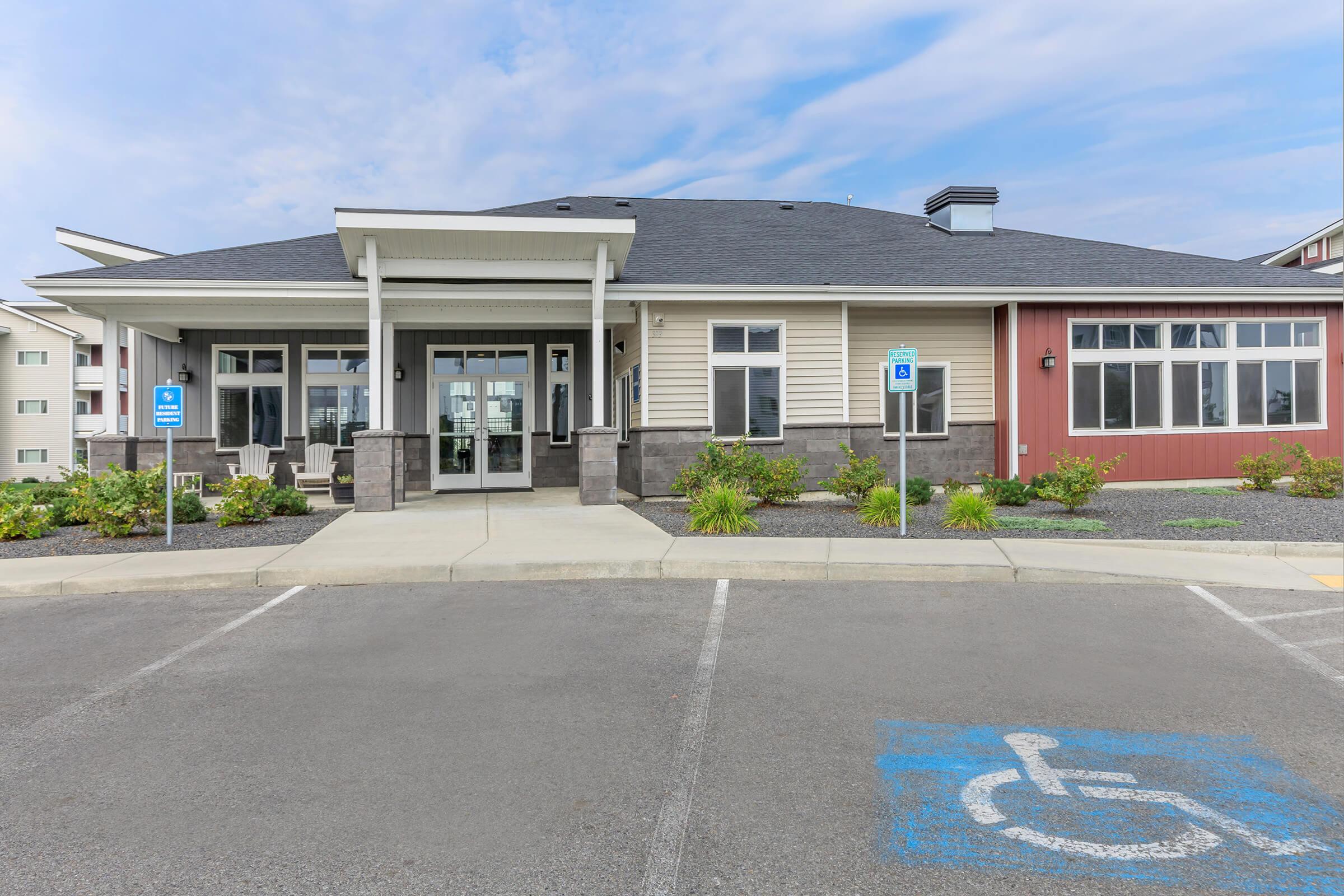 A modern one-story building featuring a combination of light and dark siding with large windows. The entrance has covered awning and is flanked by landscaped areas. In front, there are designated parking spaces, including accessible parking. The sky is partly cloudy.