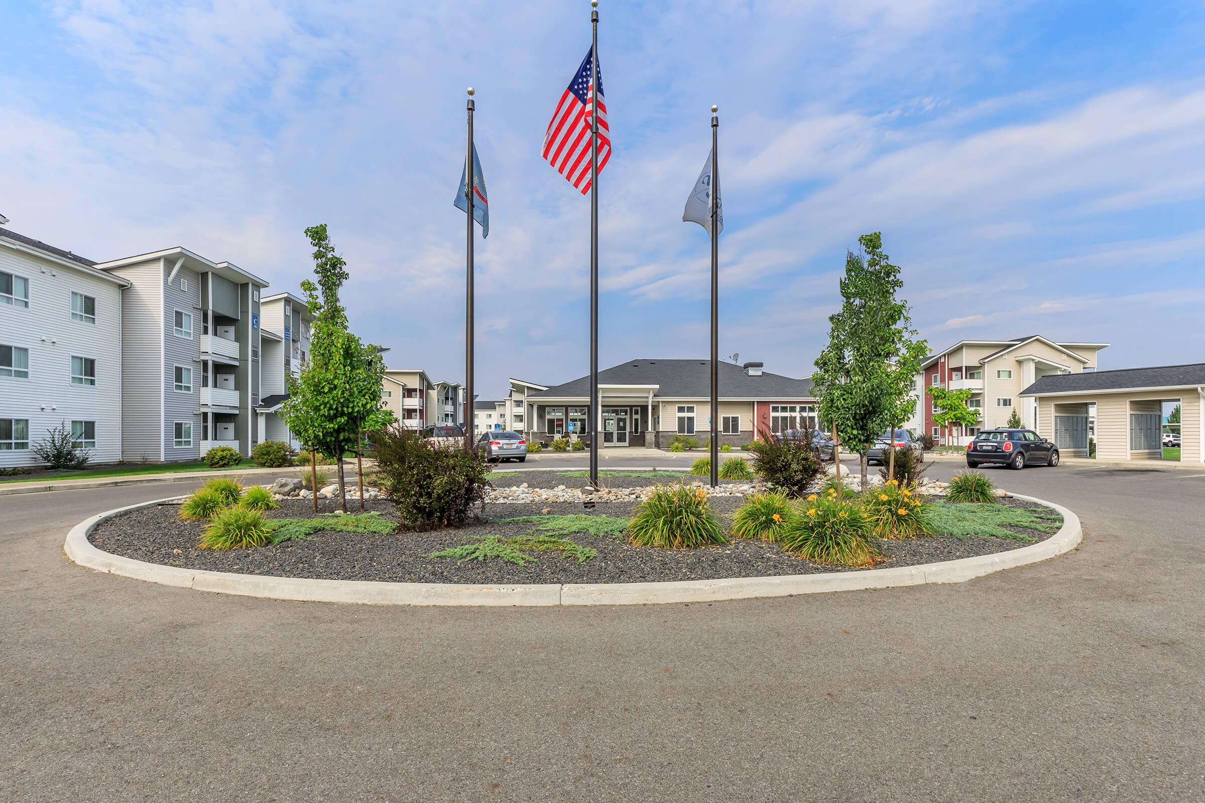 A well-maintained apartment complex entrance featuring a circular driveway. In the center, three flag poles display the American flag and two other flags. Surrounding the driveway are landscaped areas with shrubs and flowers, and modern buildings visible on either side. The sky is mostly clear with soft clouds.