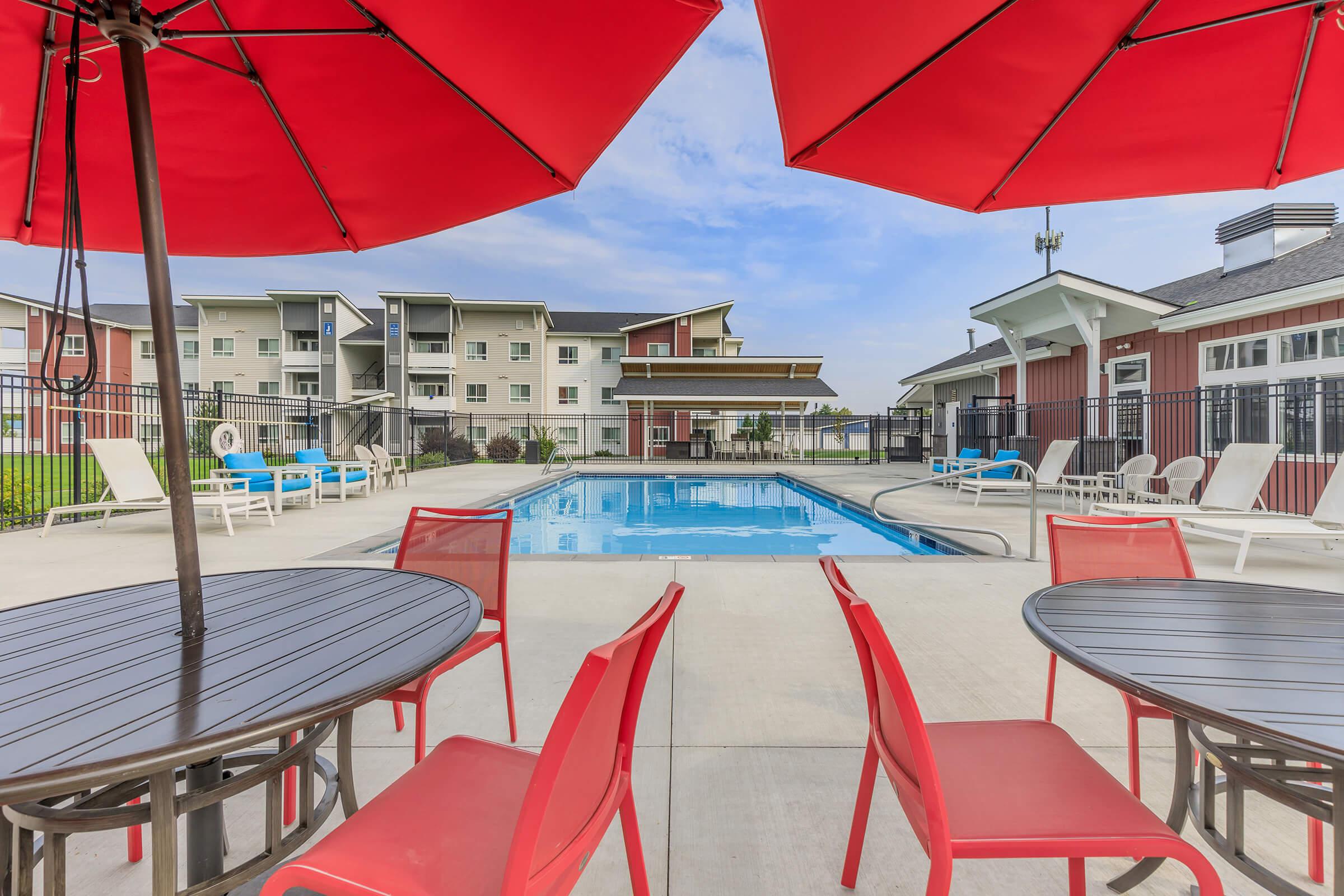 View of a swimming pool surrounded by lounge chairs and tables with red umbrellas. In the background, there are modern apartment buildings and a clear blue sky. The setting is inviting and perfect for relaxation.