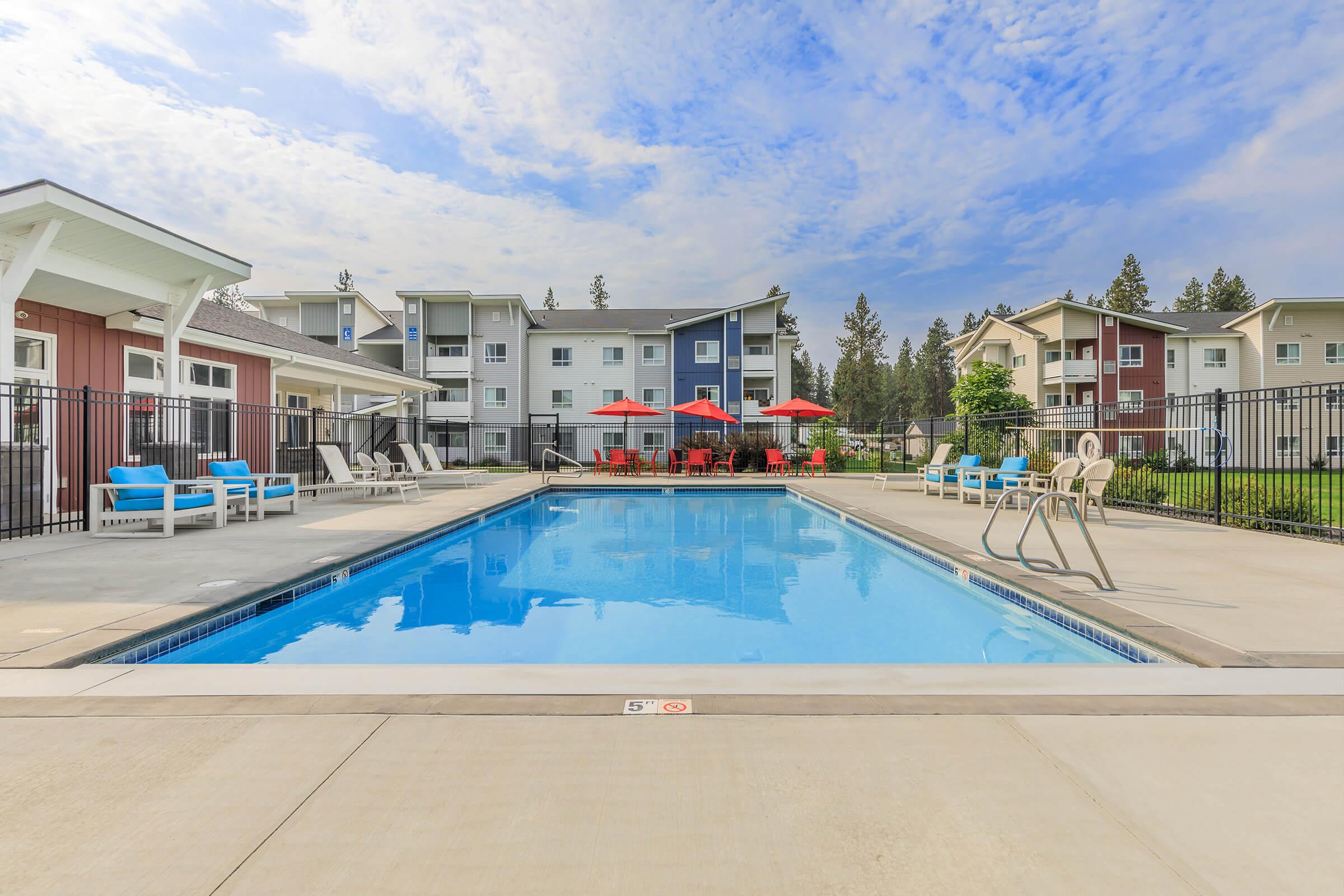A swimming pool surrounded by lounge chairs and tables with umbrellas, set in a residential complex. In the background, two-story apartment buildings are visible under a partly cloudy sky. The pool area is fenced for safety and has a clear view of the surrounding landscape.