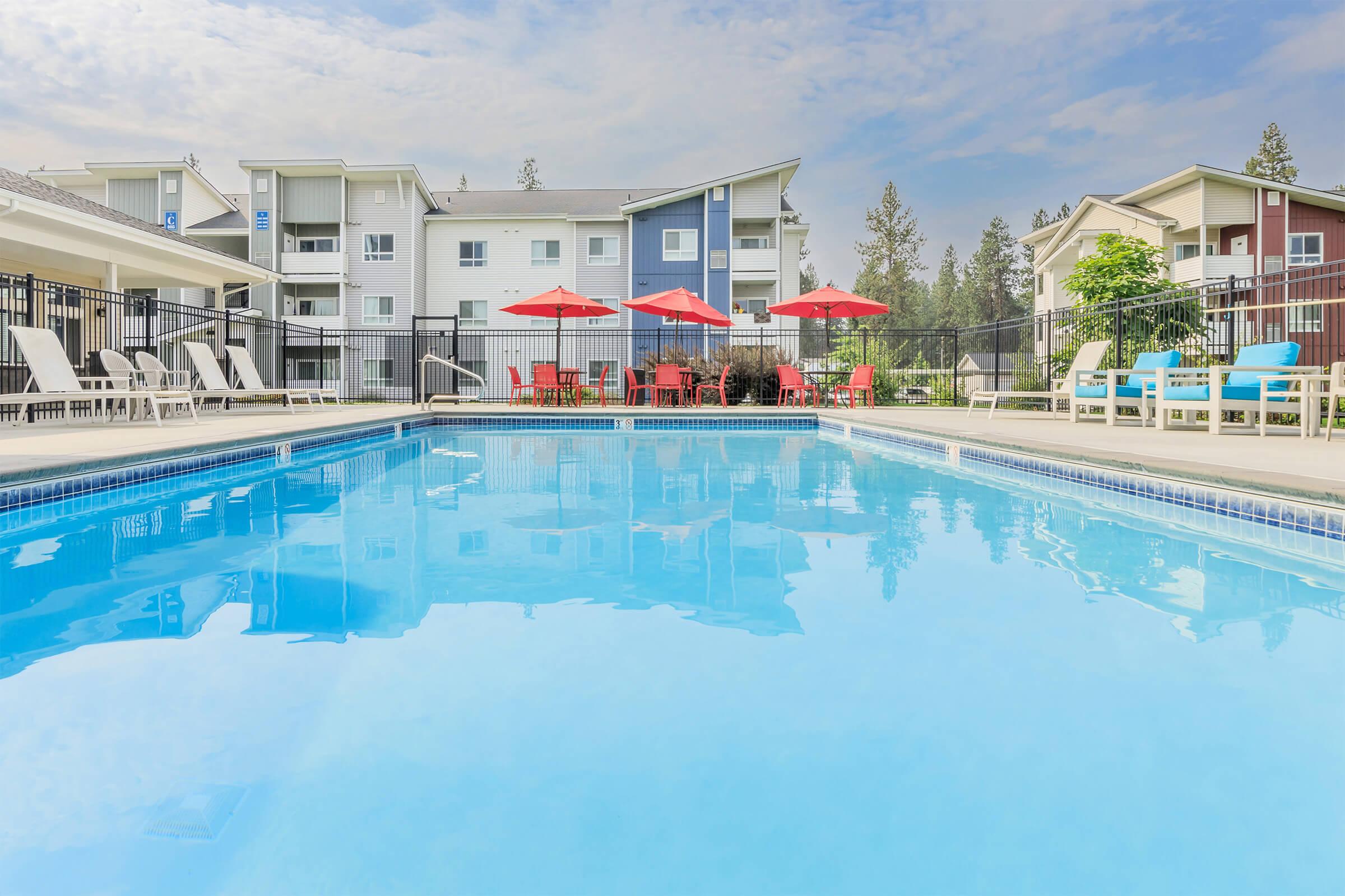 A clear blue swimming pool with lounge chairs and red umbrellas around it, surrounded by a landscaped area and modern residential buildings in the background under a cloudy sky.